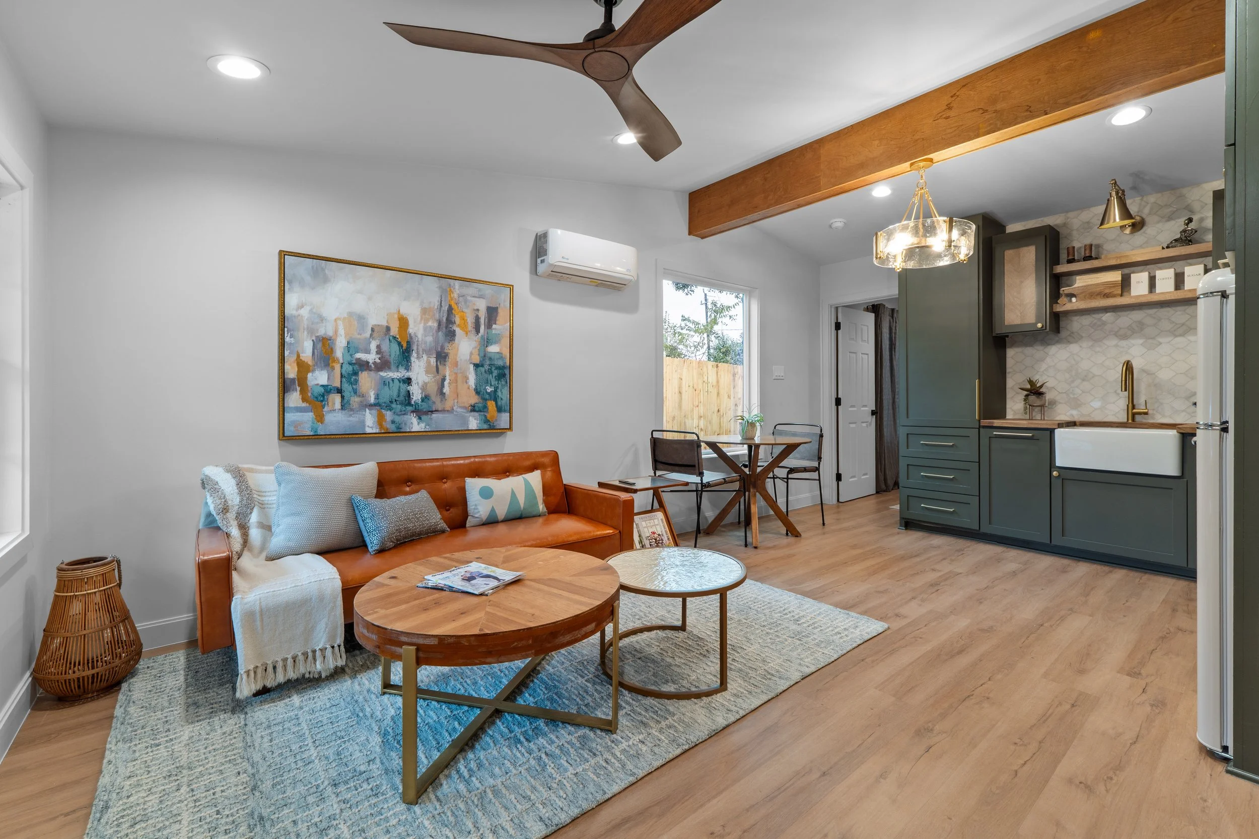 Modern living room and kitchen with white walls, wooden flooring, a leather sofa decorated with pillows, a round wooden coffee table, a blue area rug, a colorful abstract painting, and a kitchen area with green cabinets, open shelves, and a hanging light fixture.