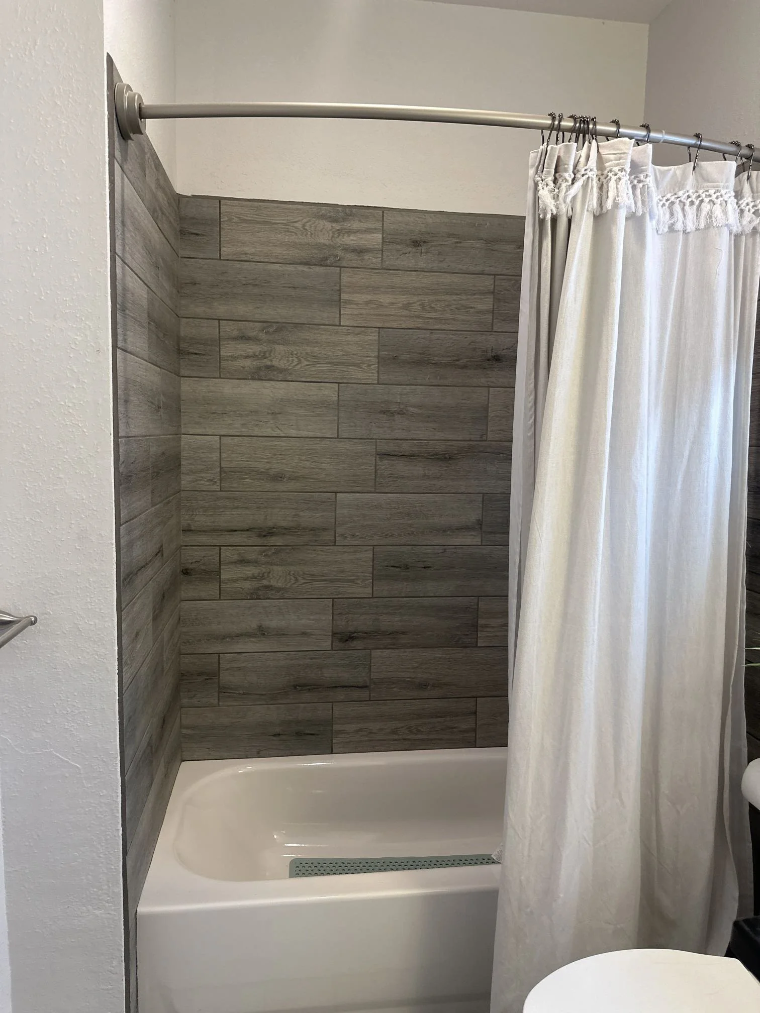 A bathroom shower area with a curved metal shower curtain rod and white curtains with fringe, wood-look tile on the wall, a white bathtub, and a small round white table in front.