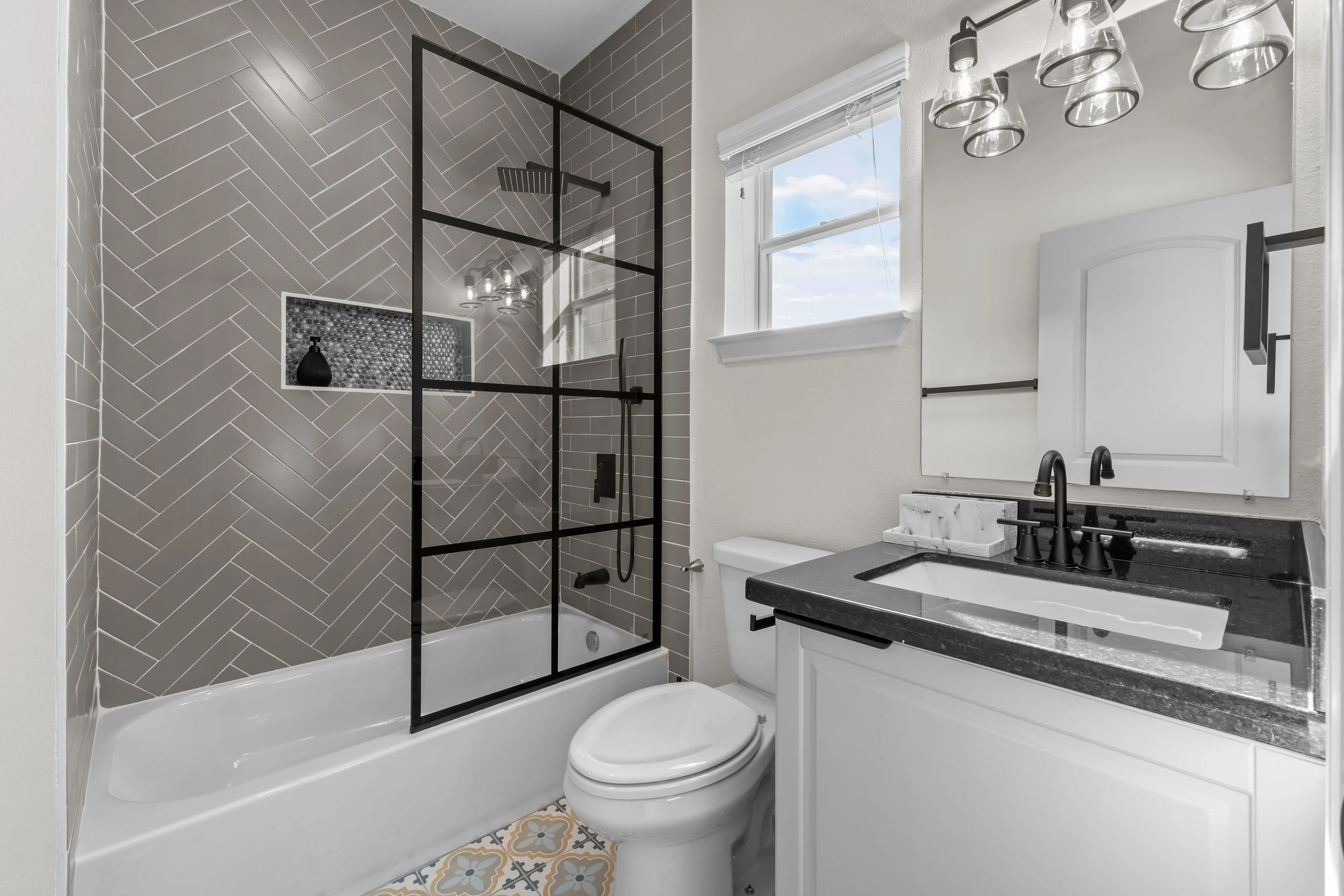 Modern bathroom with a walk-in shower featuring gray herringbone tile, black framing, and a small shelf with a black vase. There is a window with a white frame, a white toilet, and a white vanity with a black countertop and black fixtures. The floor has patterned tiles.