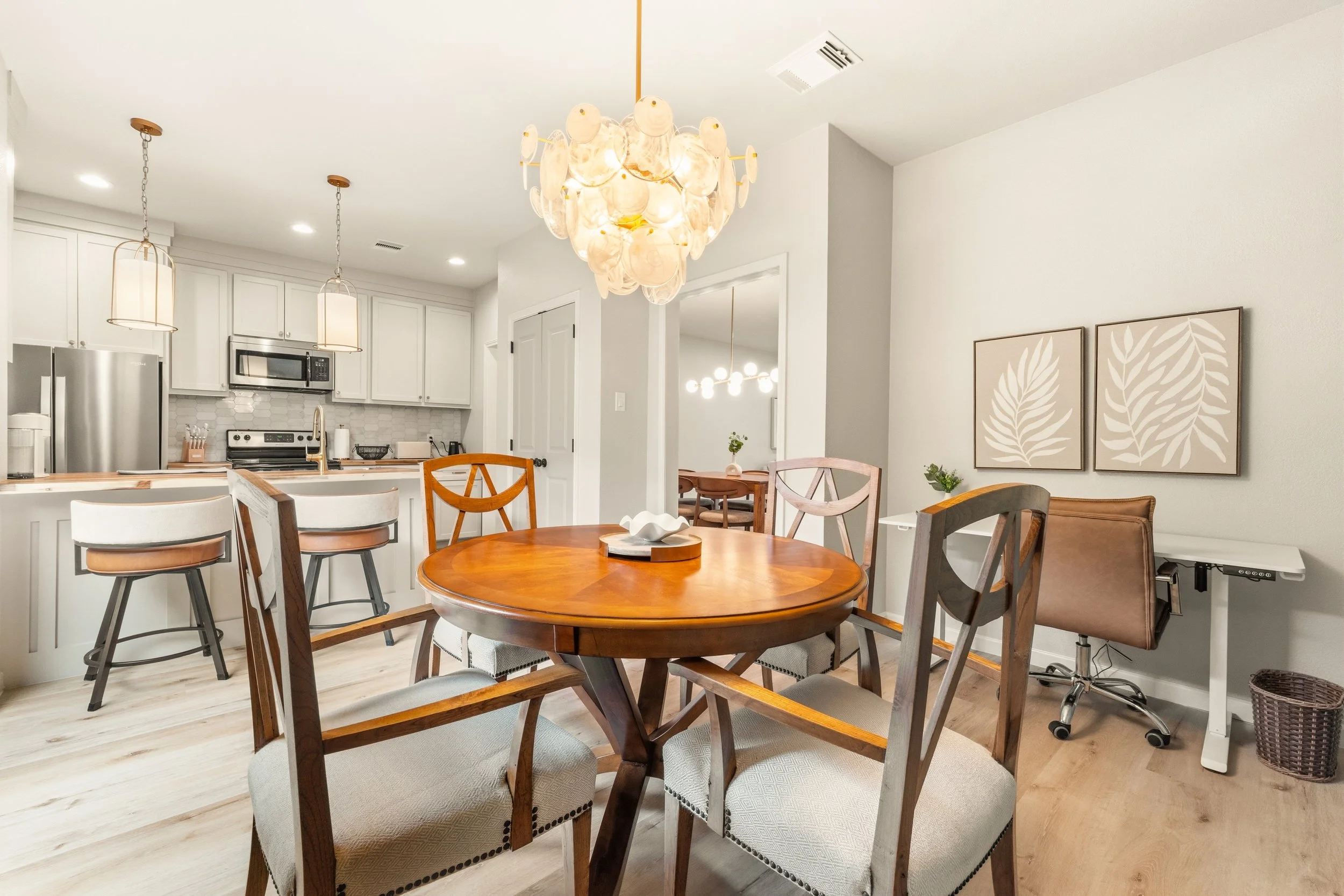 A kitchen and dining area with a round wooden table and chairs, white cabinets, stainless steel refrigerator, microwave, and a chandelier hanging from the ceiling.