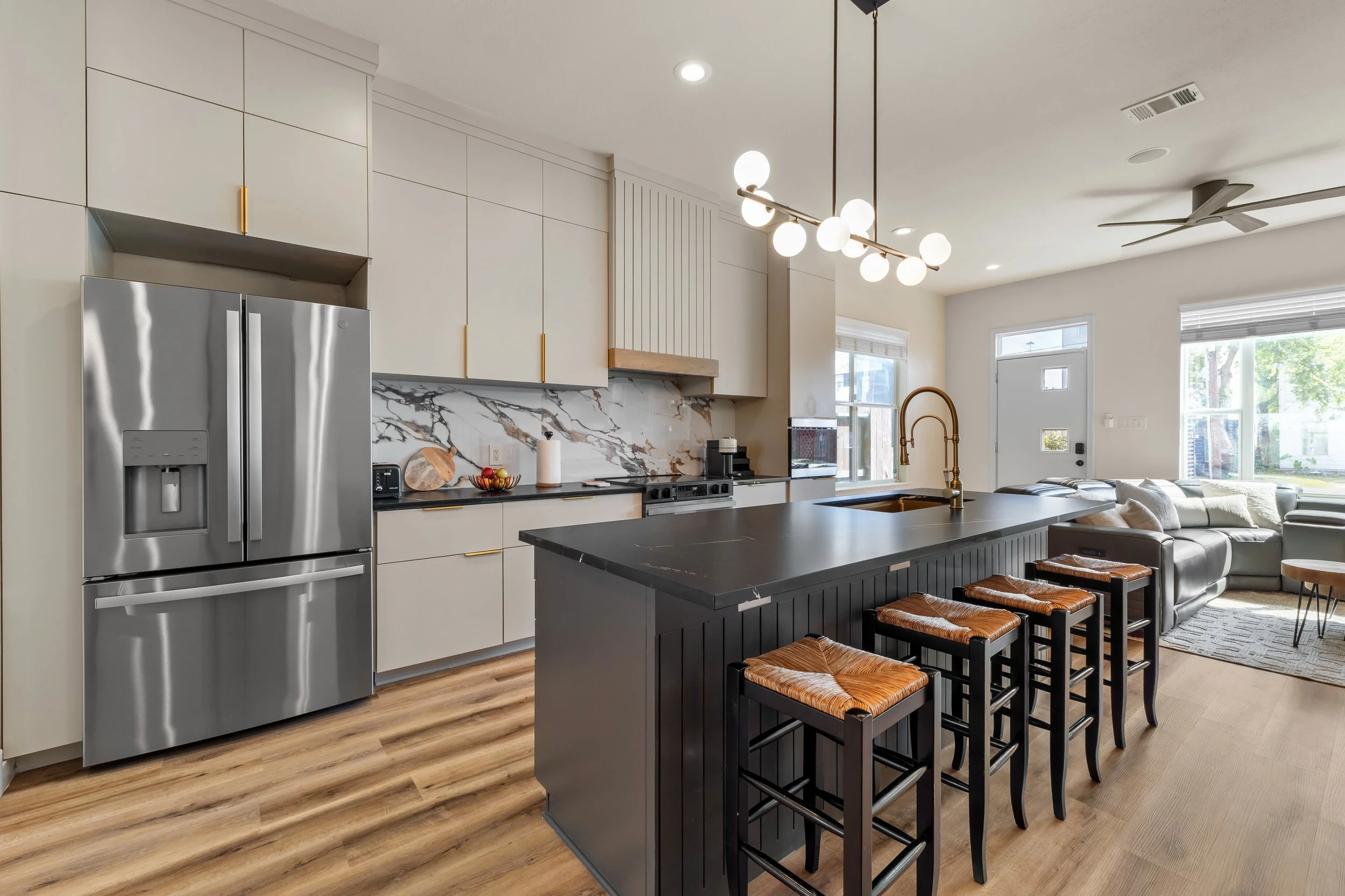 Modern kitchen with white cabinets, marble backsplash, black island with bar stools, stainless steel refrigerator, and office space near windows with bright sunlight
