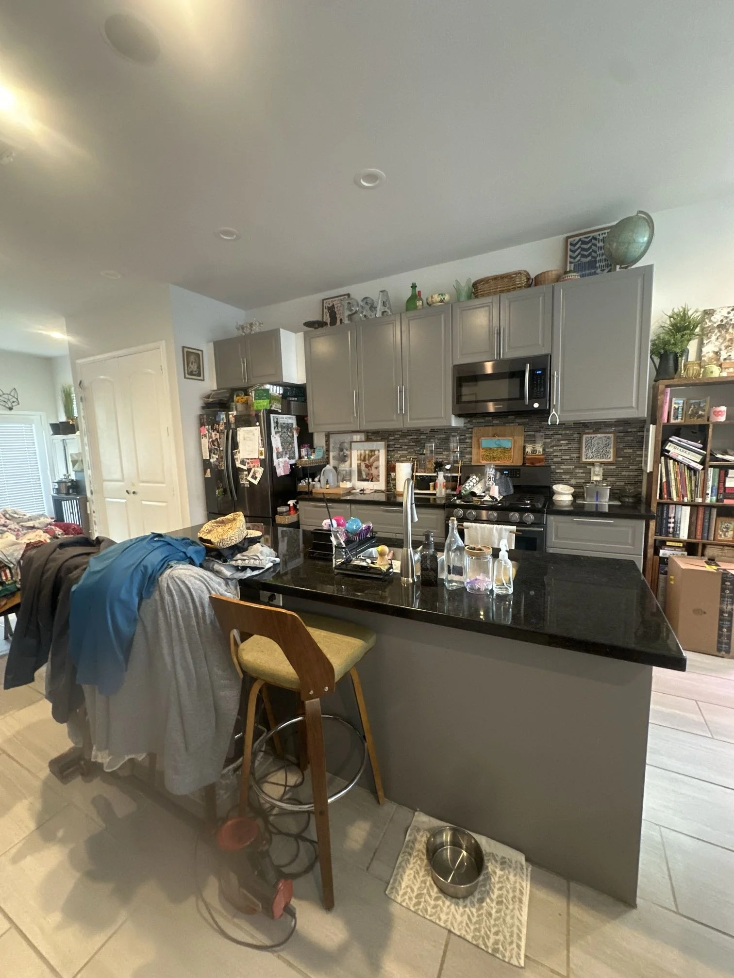 Kitchen with gray cabinets, stainless steel appliances, a black countertop, and various items on the counters and shelves, including a globe and decorative letters. There is a cluttered table with clothes and a wooden chair nearby. Dog bowls are on a patterned mat on the floor.