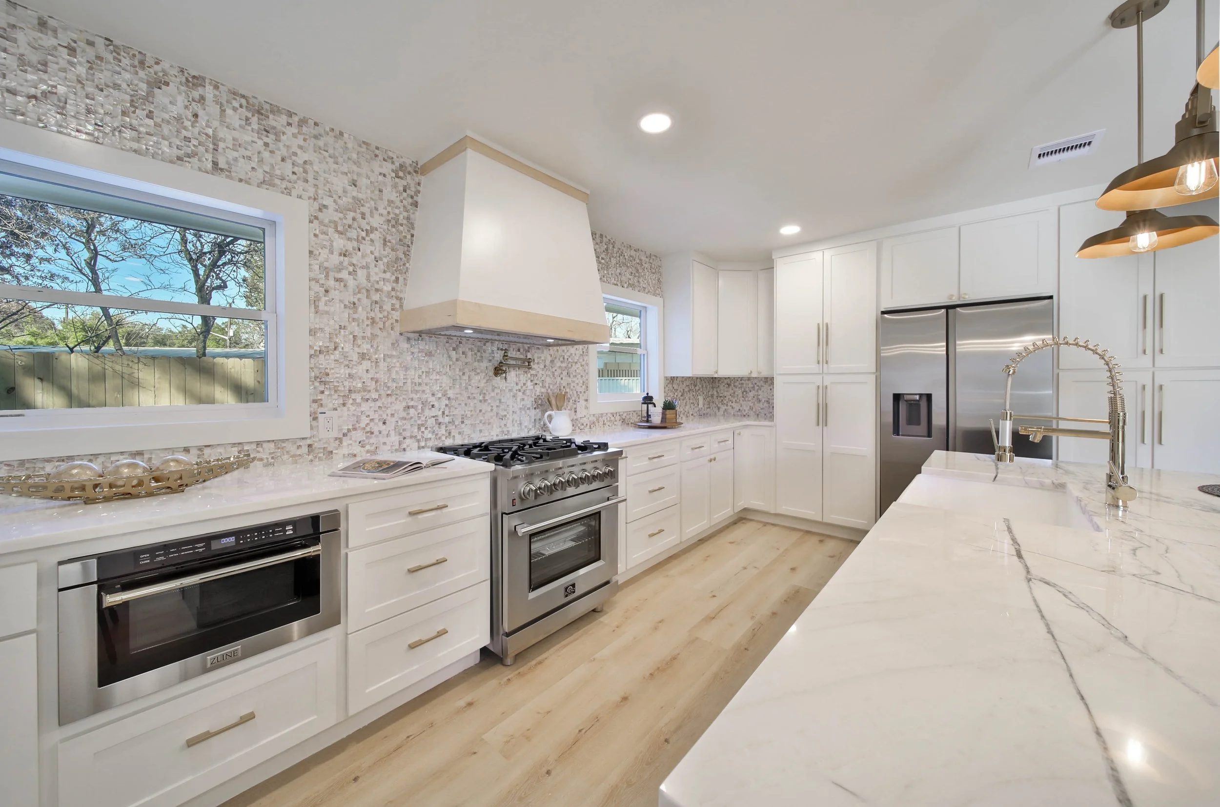 Modern white kitchen with marble countertops, stainless steel appliances, mosaic tile backsplash, large window, and pendant lighting.