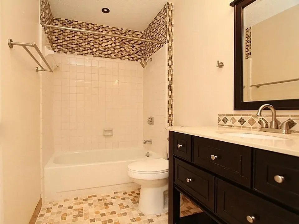 Bathroom with a bathtub and shower area, a toilet, and a black vanity with a mirror. Tiled walls and floor with a mosaic border.