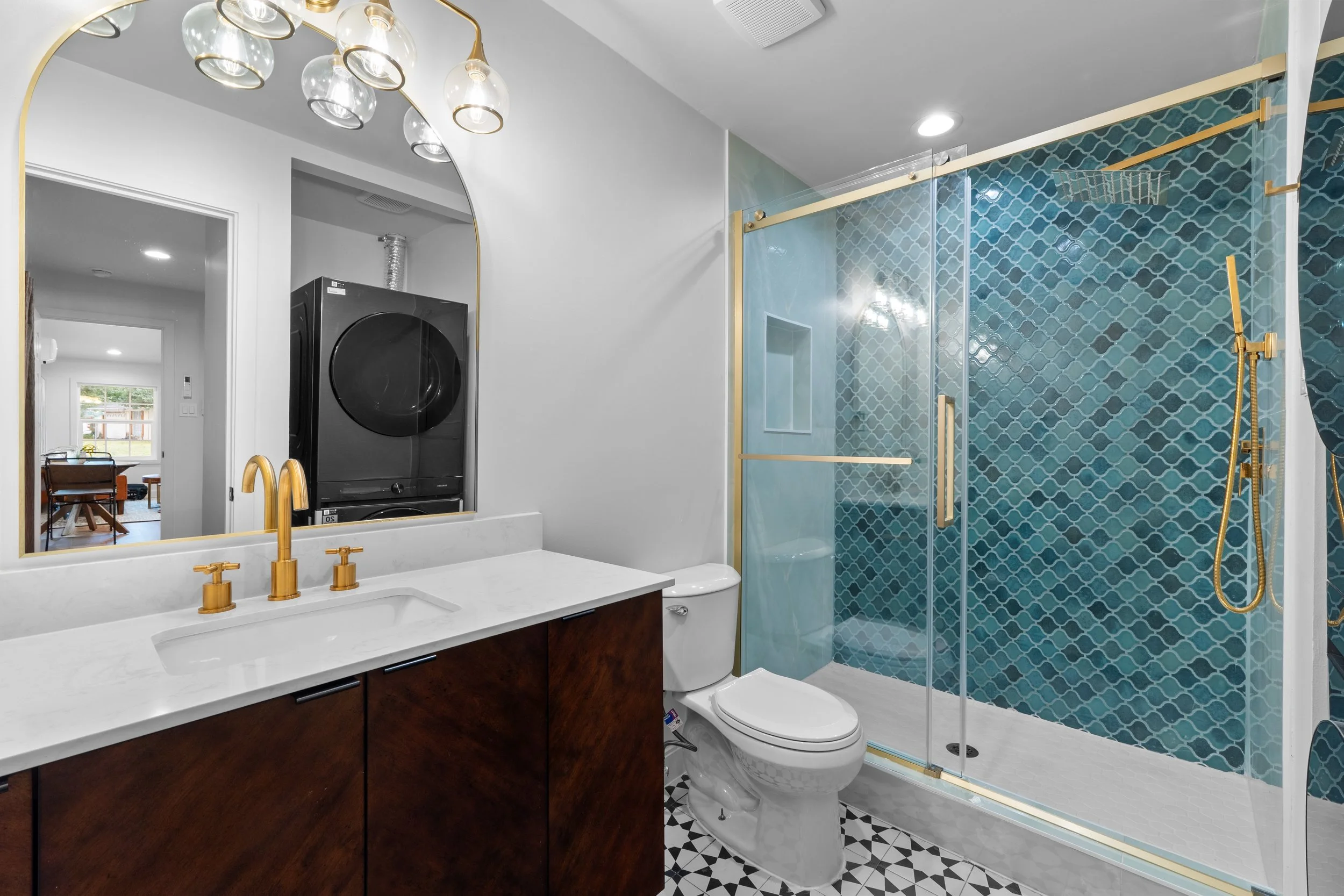 Modern bathroom with a vanity mirror, shelf, and gold fixtures, a shower with glass doors and blue-patterned tiles, a white toilet, and a black laundry machine visible through the mirror.