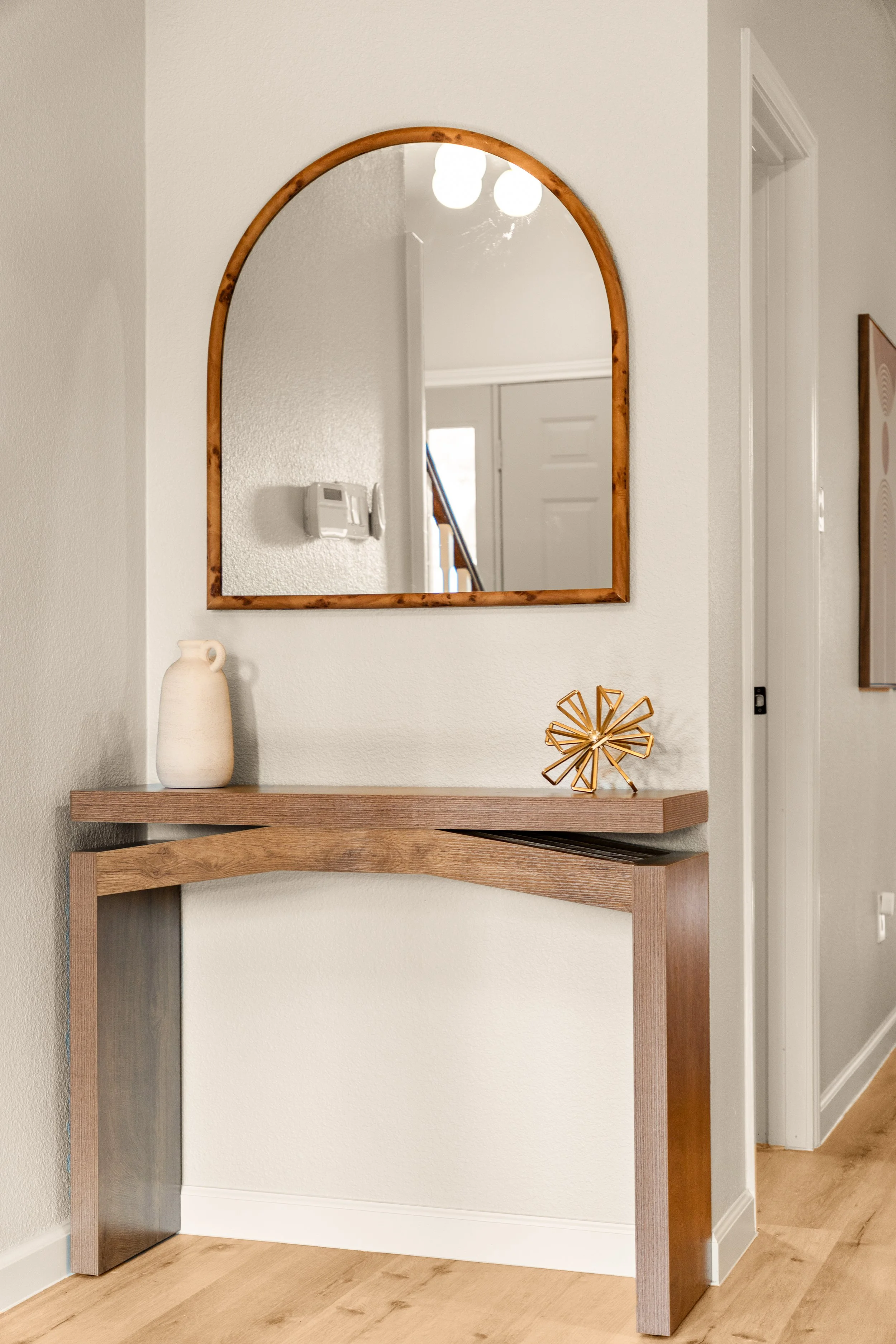 A wall mirror with a wooden frame reflecting a ceiling light fixture, positioned above a wooden floating shelf with decorative items, and adjacent to a white door and textured wall.