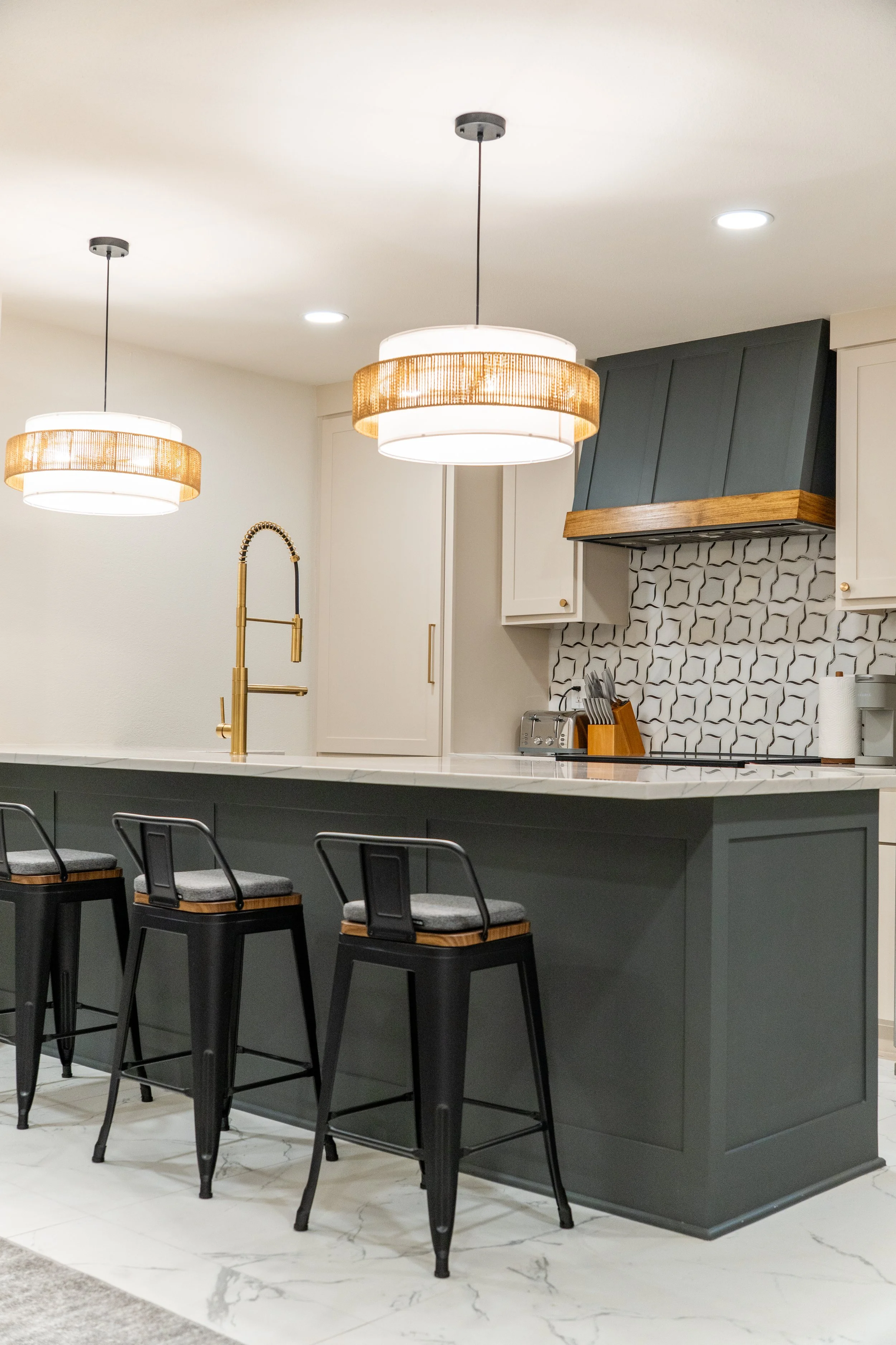 Modern kitchen with a white marble island, black bar stools, and a dark green range hood over a patterned backsplash. Two pendant lights hang above the island.