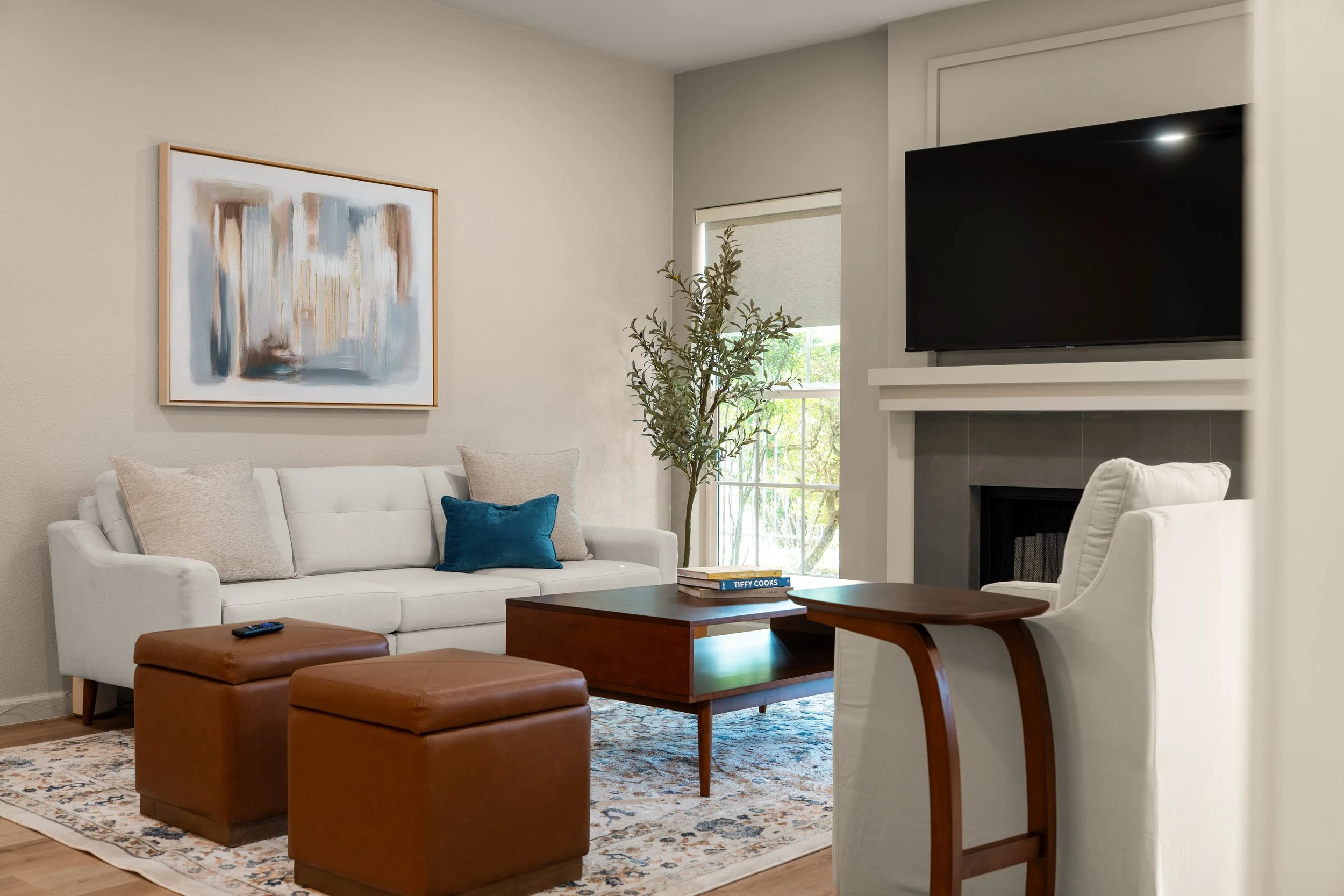 Living room with white sofa, throw pillows, wooden coffee table, ottomans, a decorative rug, wall art, a window with blinds, a tall plant, and a mounted TV above a fireplace.