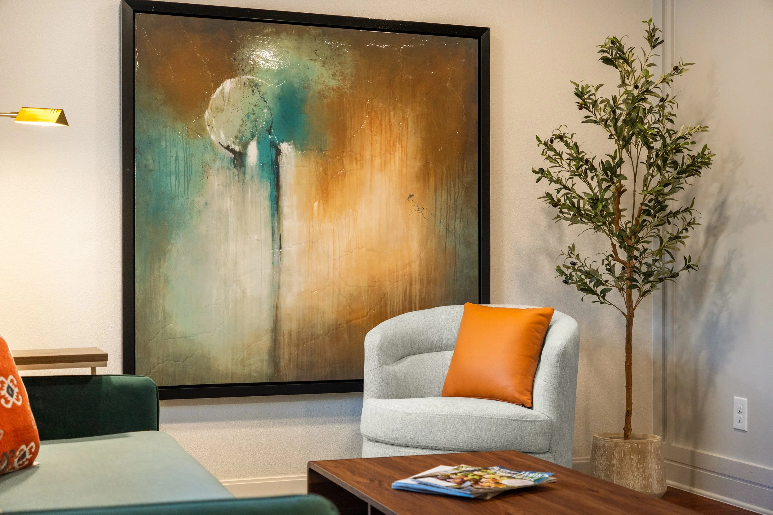 Living room with a large framed abstract painting, a white armchair with an orange pillow, a potted plant, a wooden coffee table with magazines, and partially visible seating with a patterned pillow.