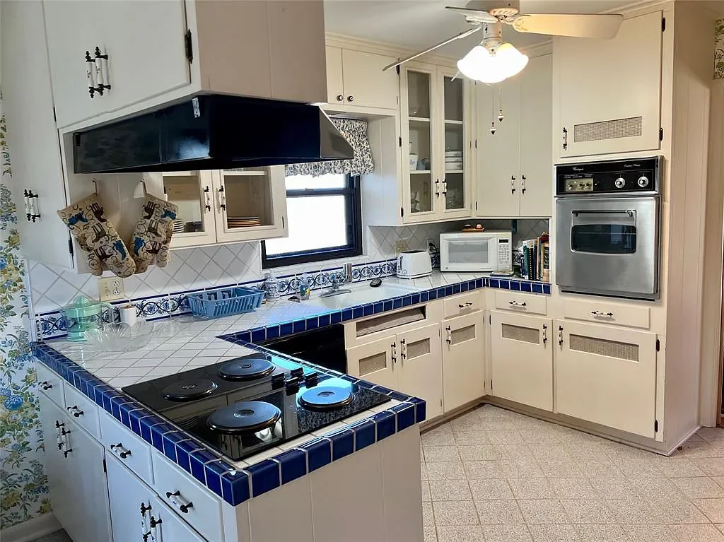 Kitchen with white cabinets, blue tiled counter edges, black electric stove, microwave, toaster, oven, and ceiling fan.