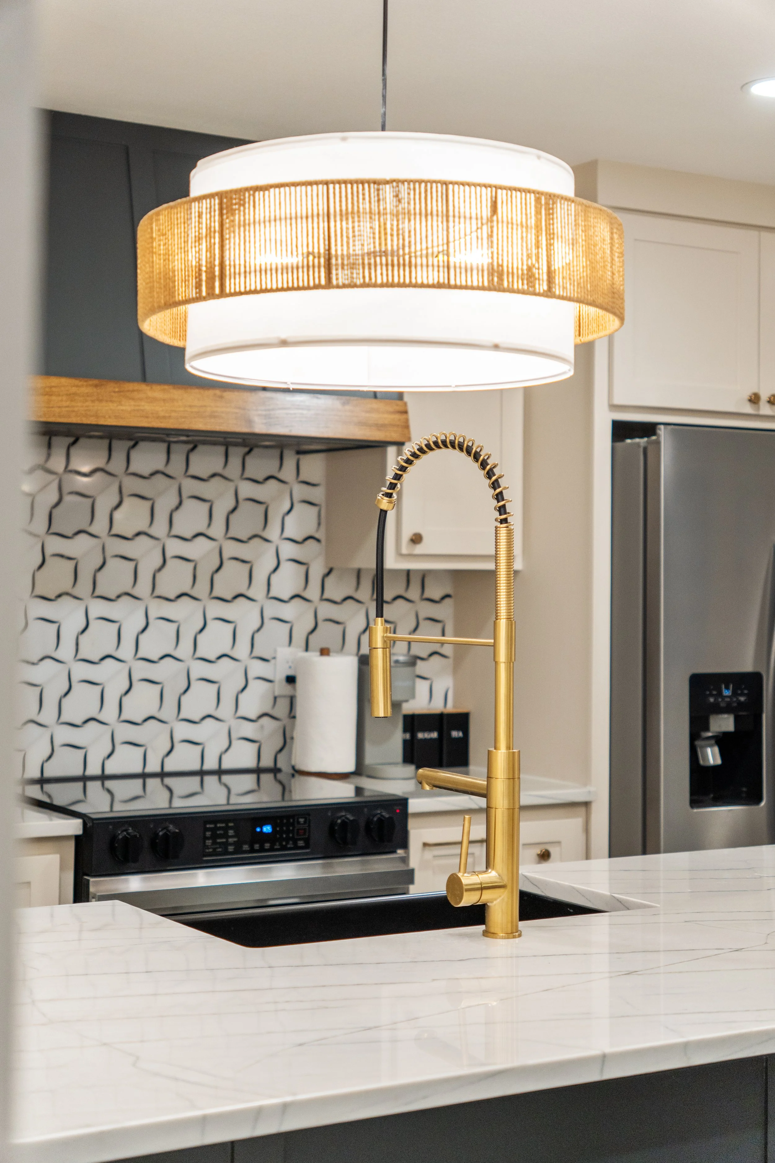 Modern kitchen with a white marble countertop, a stylish gold kitchen faucet, a black stove, a paper towel roll, and a decorative backsplash with black geometric patterns, illuminated by a large, layered ceiling light with a gold inner ring.