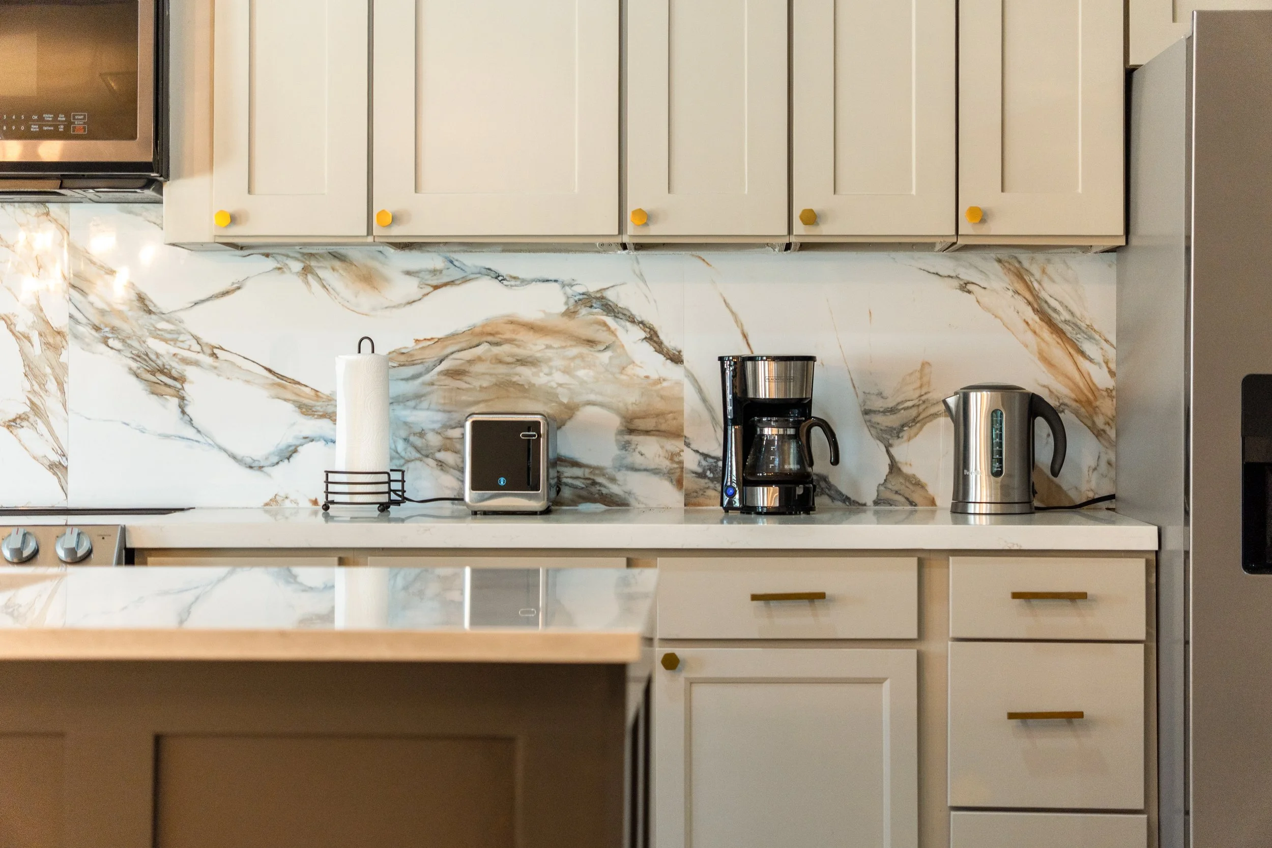 Kitchen countertop with paper towel roll, small black toaster, coffee maker, electric kettle, and marble backsplash, with white cabinets above.