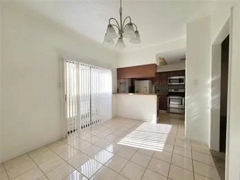 Empty living room with tiled floor, sliding glass door with vertical blinds, and a kitchen in the background.