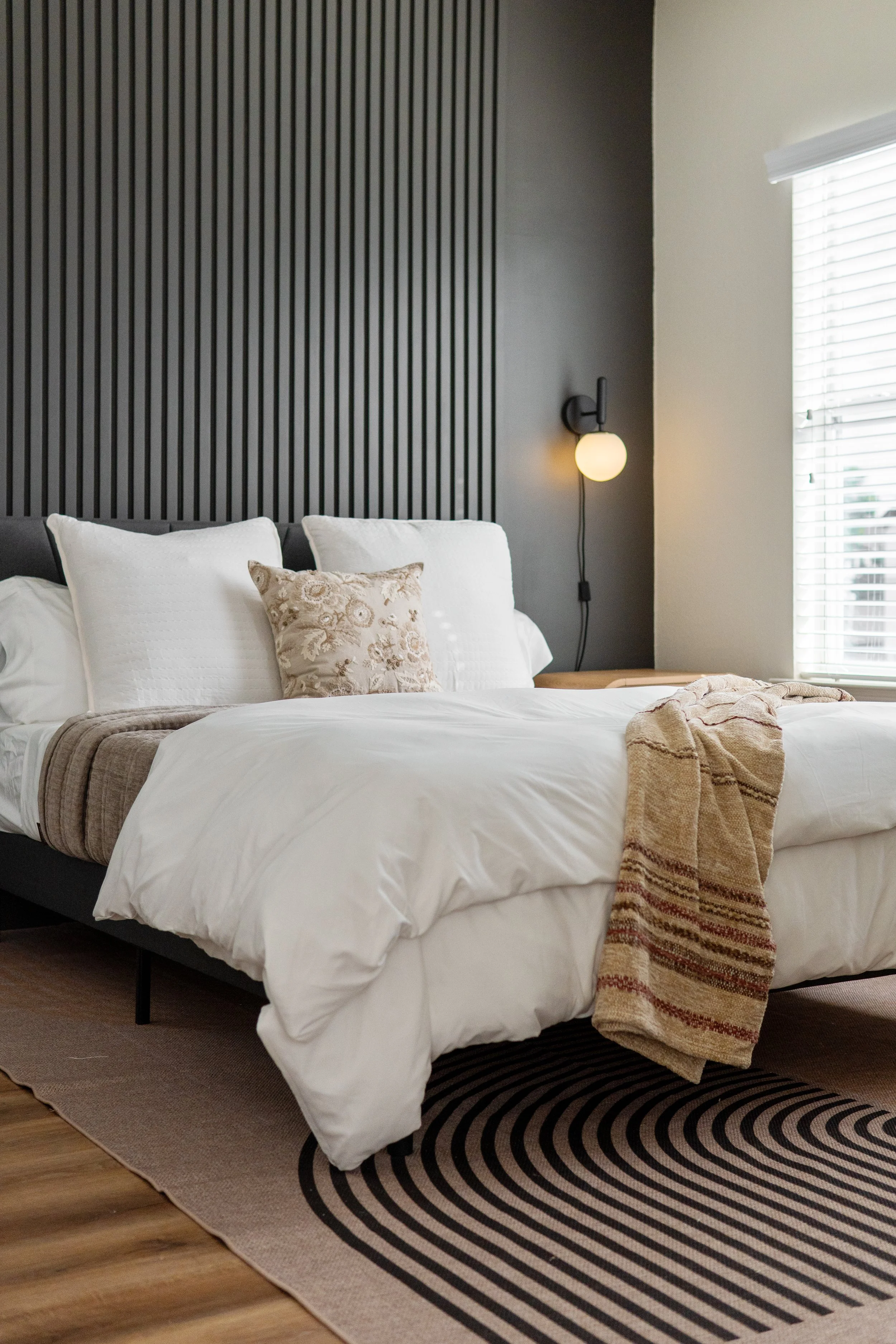 A bedroom with a bed that has white bedding and decorative pillows, a black and beige striped rug, a textured beige throw blanket, a black headboard, a dark gray wall with a round wall sconce, and a window with white blinds.