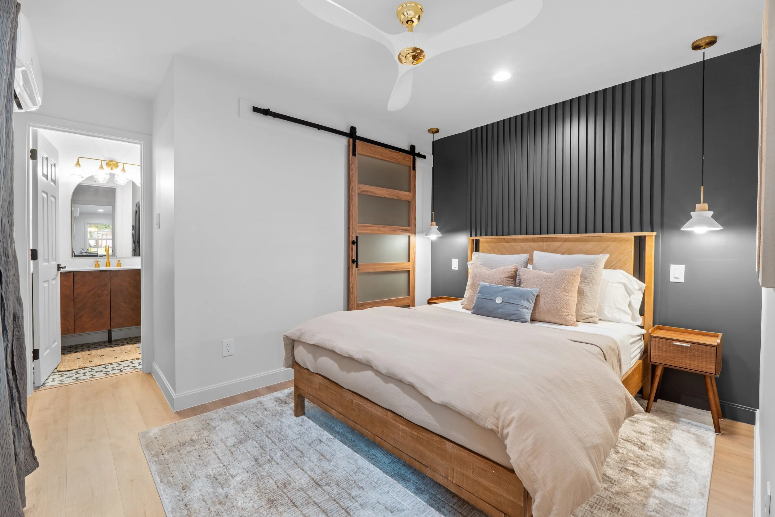 A modern bedroom with a light-colored bed, wooden headboard, and side table. The room features a black accent wall with wooden paneling, pendant lighting, and a ceiling fan. There is a sliding barn door and a view into a bathroom with a vanity and mirror.