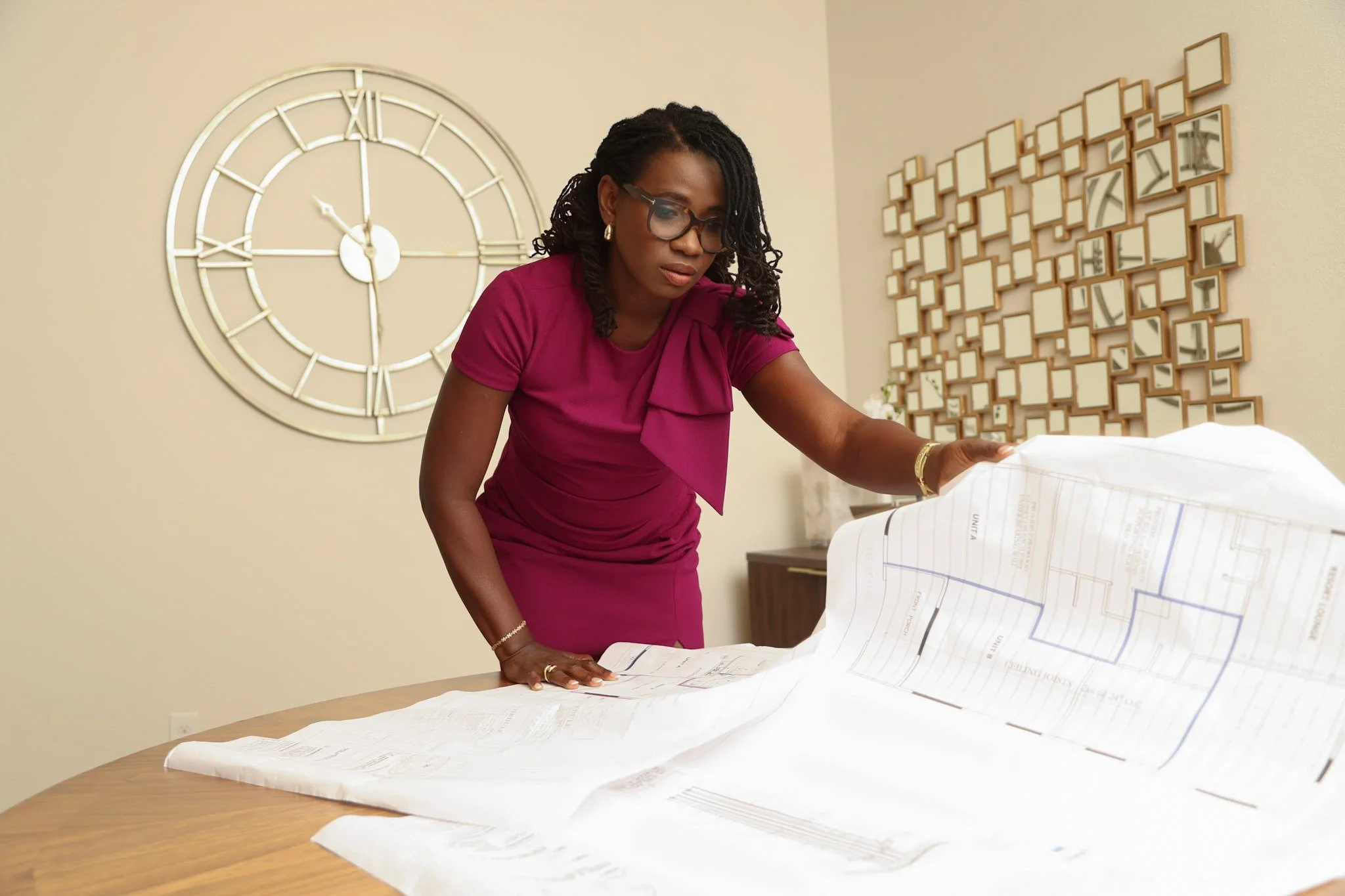 A woman in a magenta dress and glasses stands over large architectural blueprints spread across a wooden table, with a modern wall clock and a decorative mirror collage in the background.