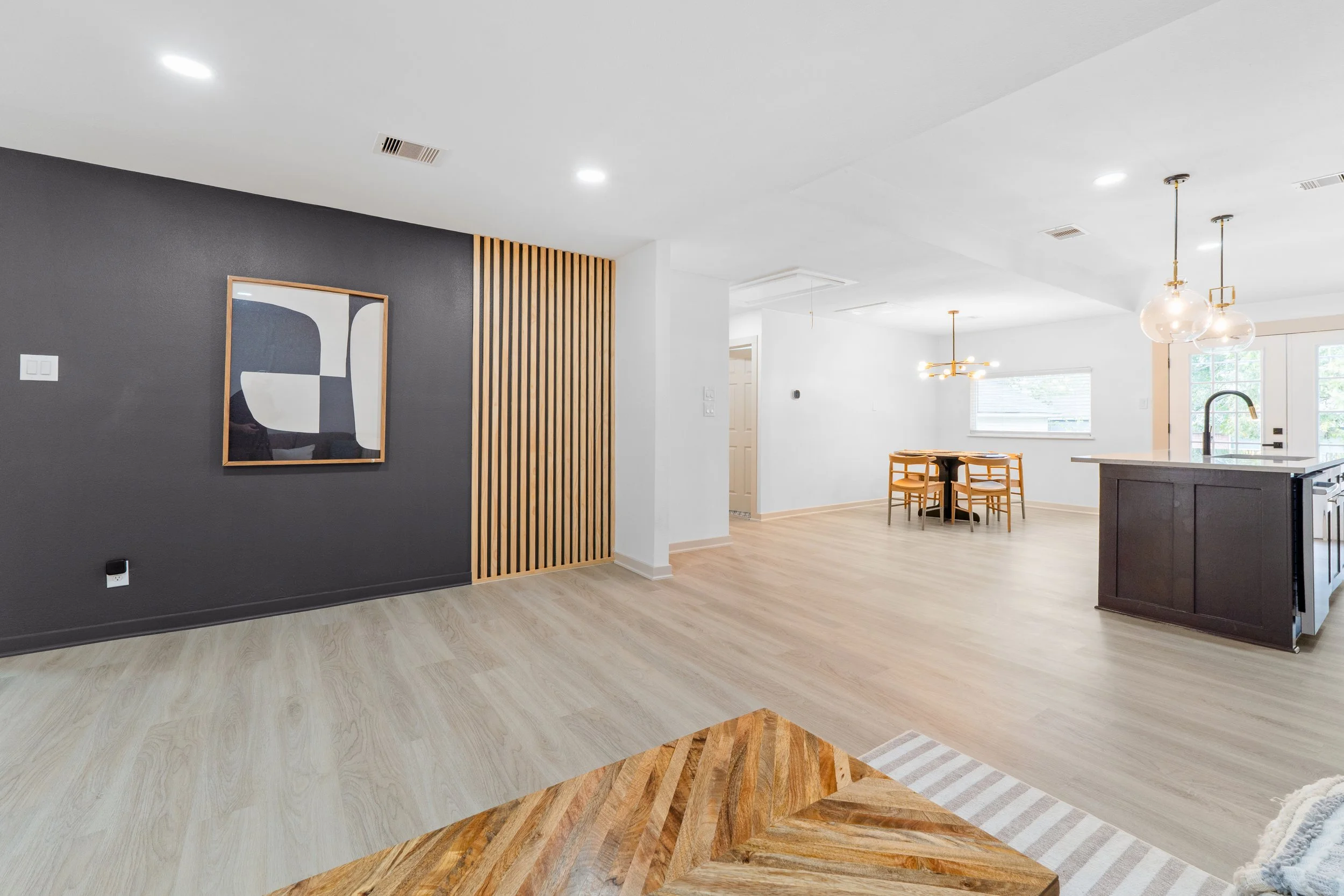 Open floor plan of a modern kitchen and dining area with white walls, large windows, and a white ceiling with recessed lighting. There's a black kitchen island with a sink, a dining table with four chairs, and pendant lights hanging above. The room has hardwood flooring and minimalist decor.
