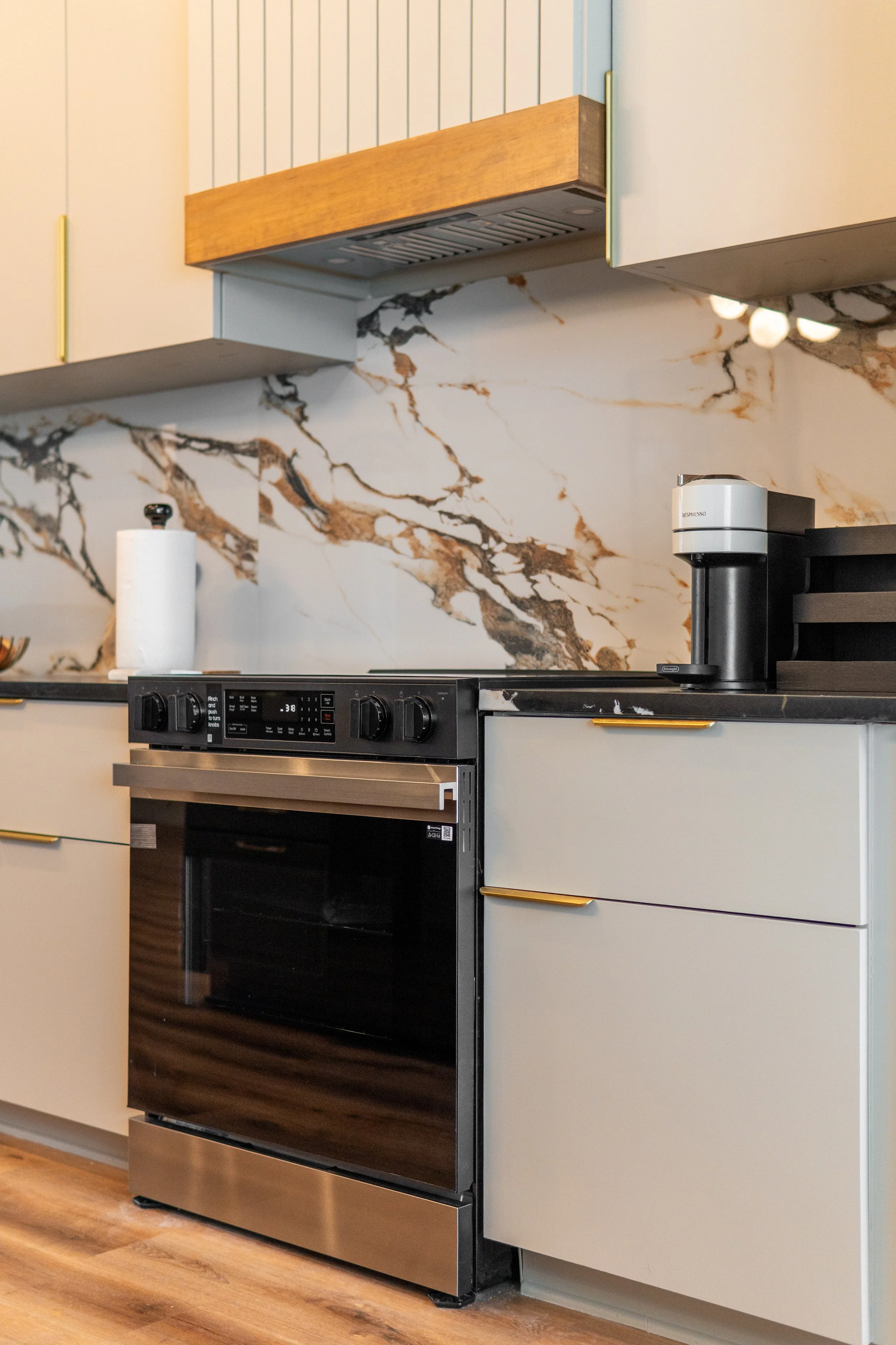 A modern kitchen with a black and stainless steel oven, white cabinets with gold handles, a marble-like backsplash, a coffee maker, and a roll of paper towels on the counter.
