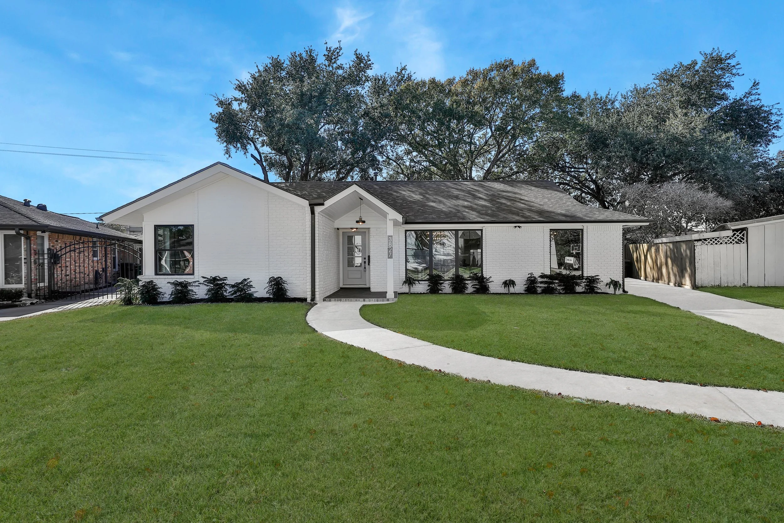 A modern single-story white brick house with a curved concrete walkway leading to the front door, surrounded by a well-manicured green lawn and a row of small bushes, with large trees in the background and a bright blue sky overhead.
