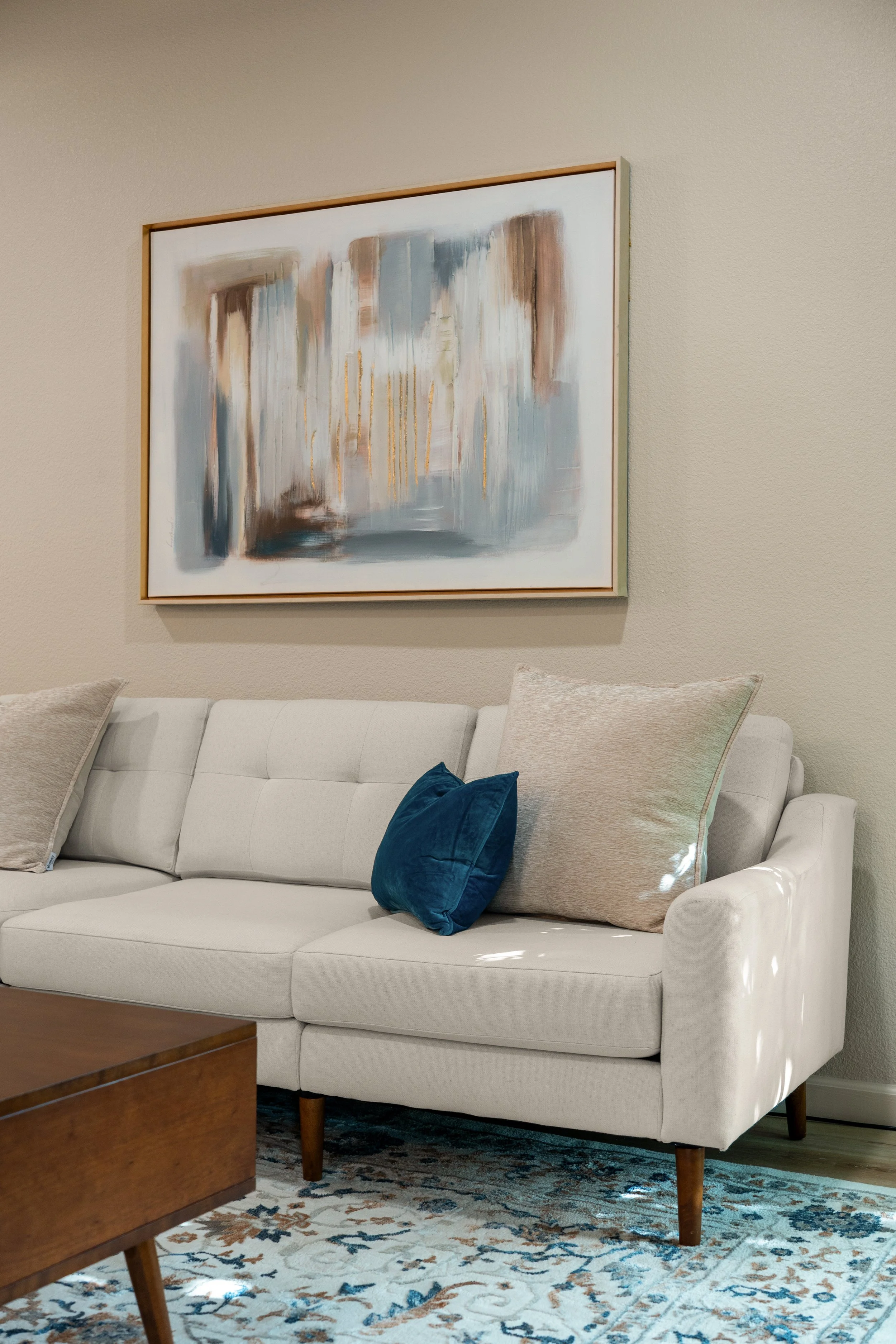 A white upholstered sofa with beige and teal throw pillows in a living room with an abstract painting on the wall, a wooden coffee table, and a patterned area rug.