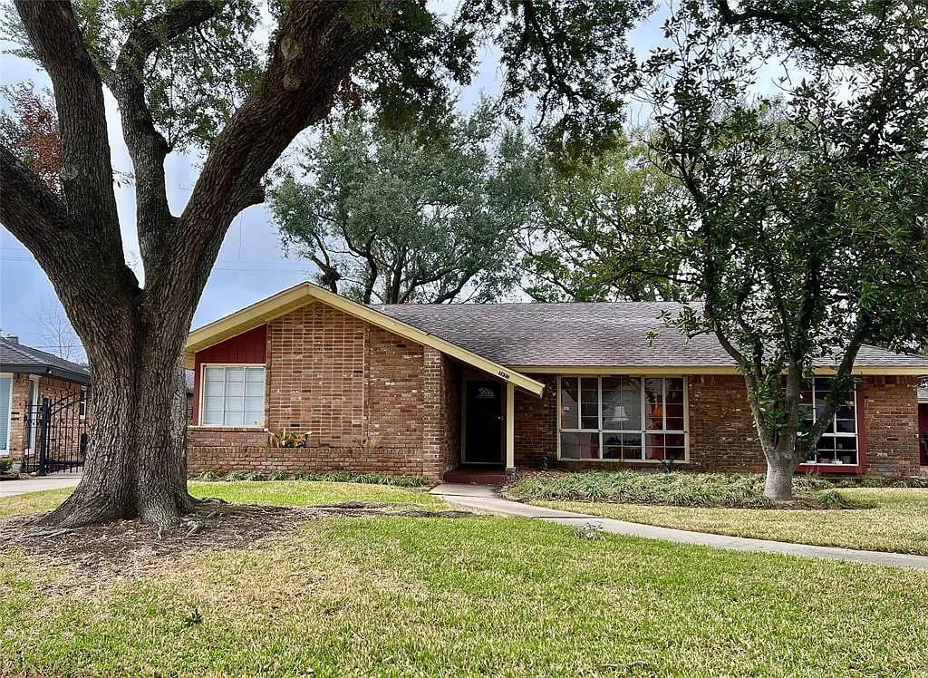 A single-story brick house with a gable roof, front yard with grass, sidewalk leading to the front door, and trees in the yard.