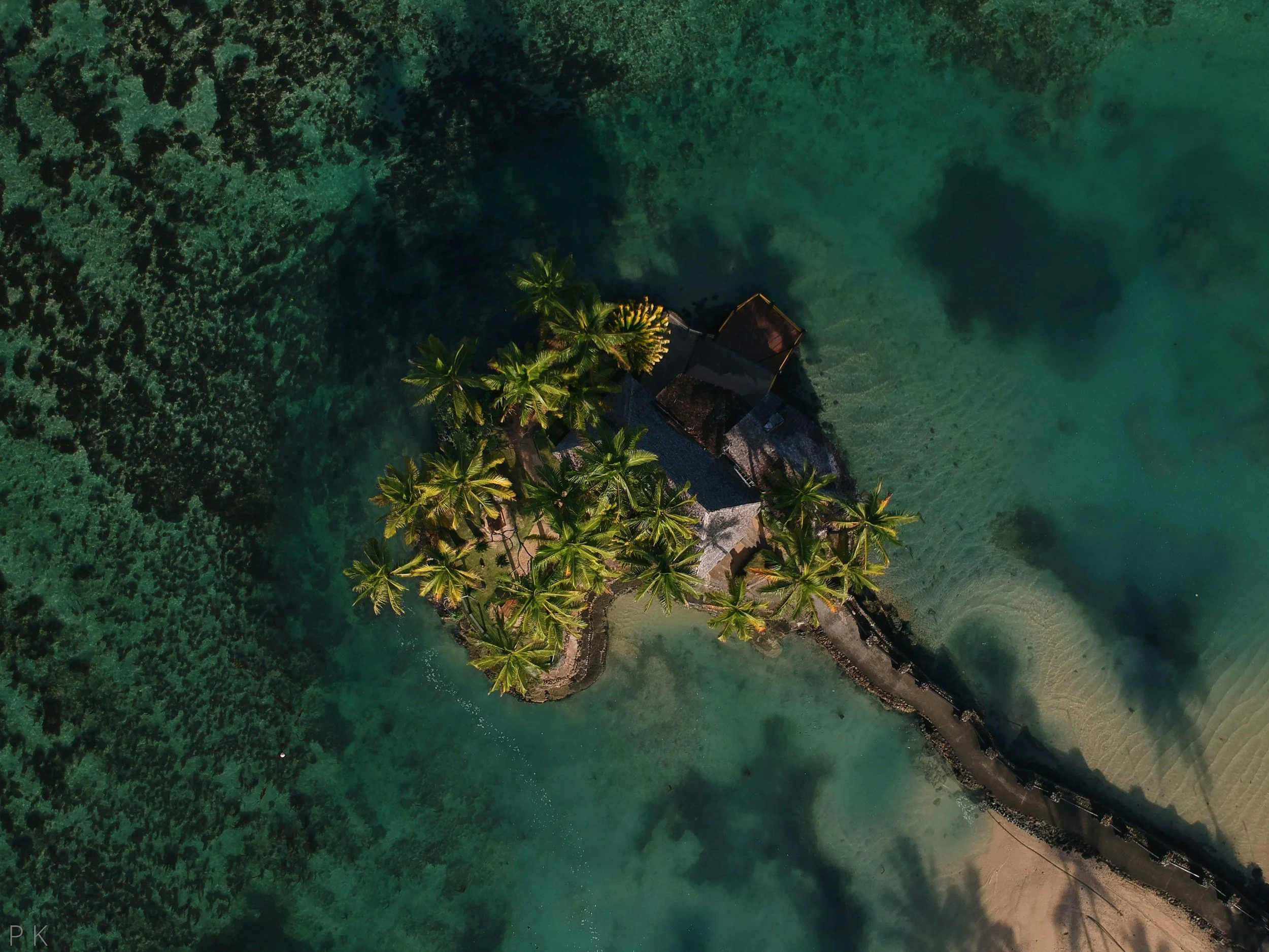 Aerial view of a small island with palm trees, surrounded by clear turquoise water, with a narrow strip of shoreline.