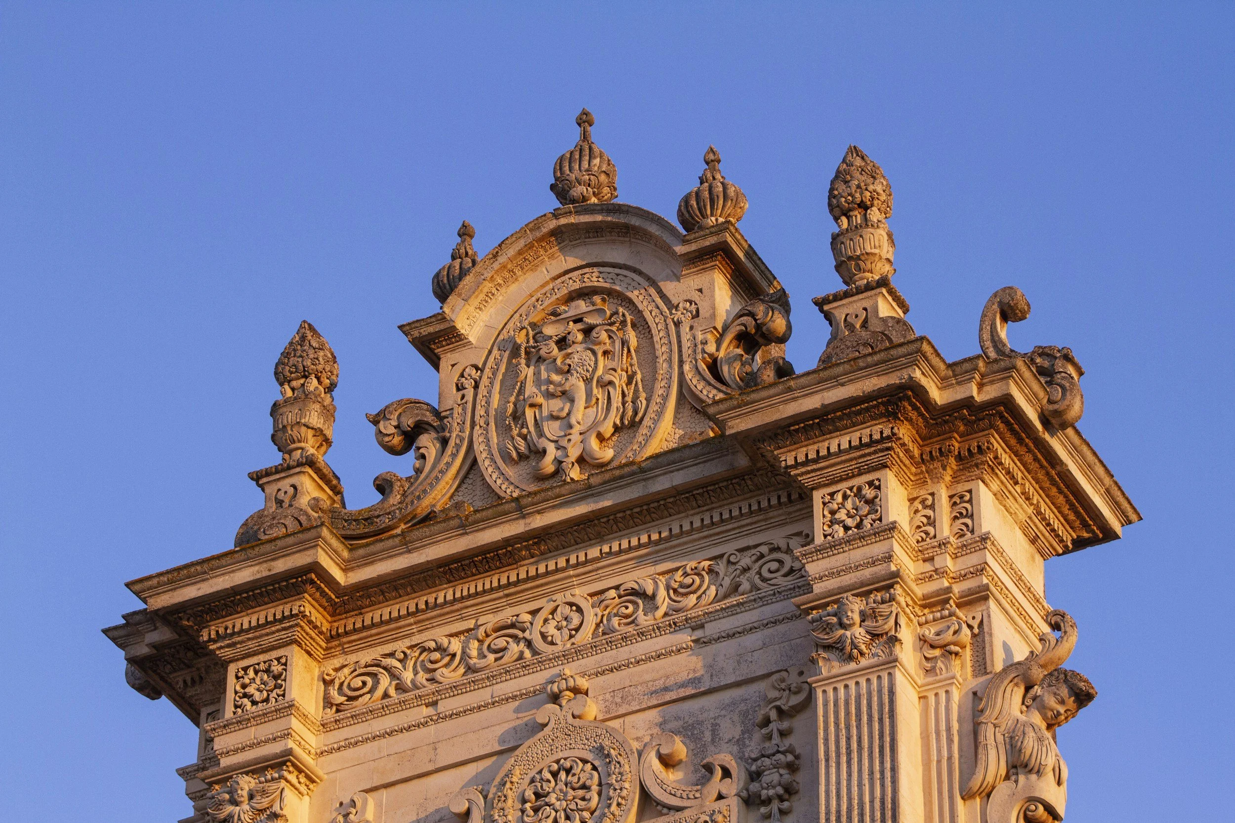 Close-up of ornate historic building with intricate stone carvings and decorative elements, against a clear blue sky. Lecce, Puglia Italy.