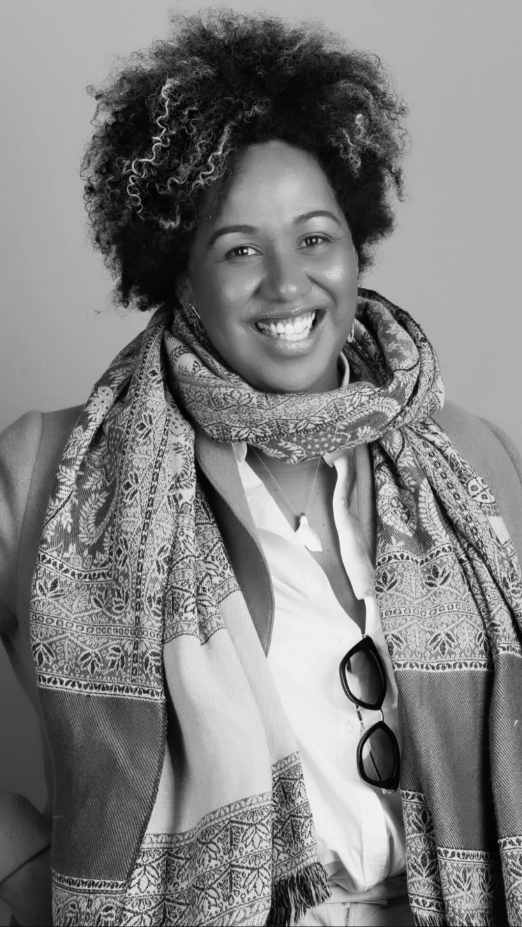 Black and white portrait of a smiling woman with curly hair, wearing a patterned scarf, glasses hanging from her shirt, and a blazer. Elle McCarthy, Kalokalo travel