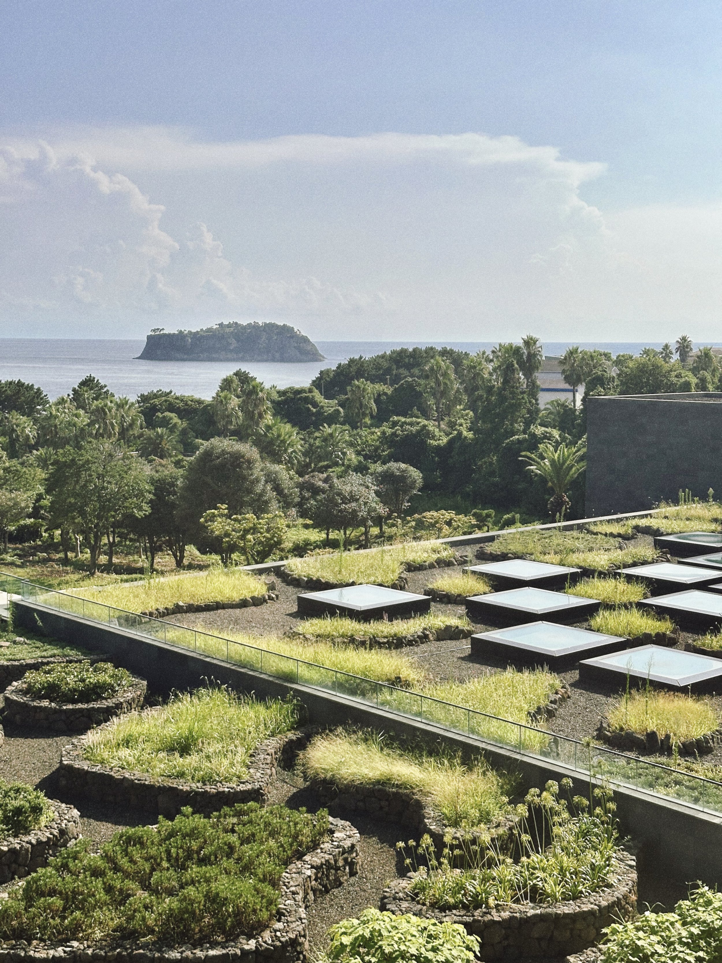 A landscape view of a garden with various green plants and trees, overlooking a body of water with an island in the distance, under a partly cloudy sky. Marriott Jeju