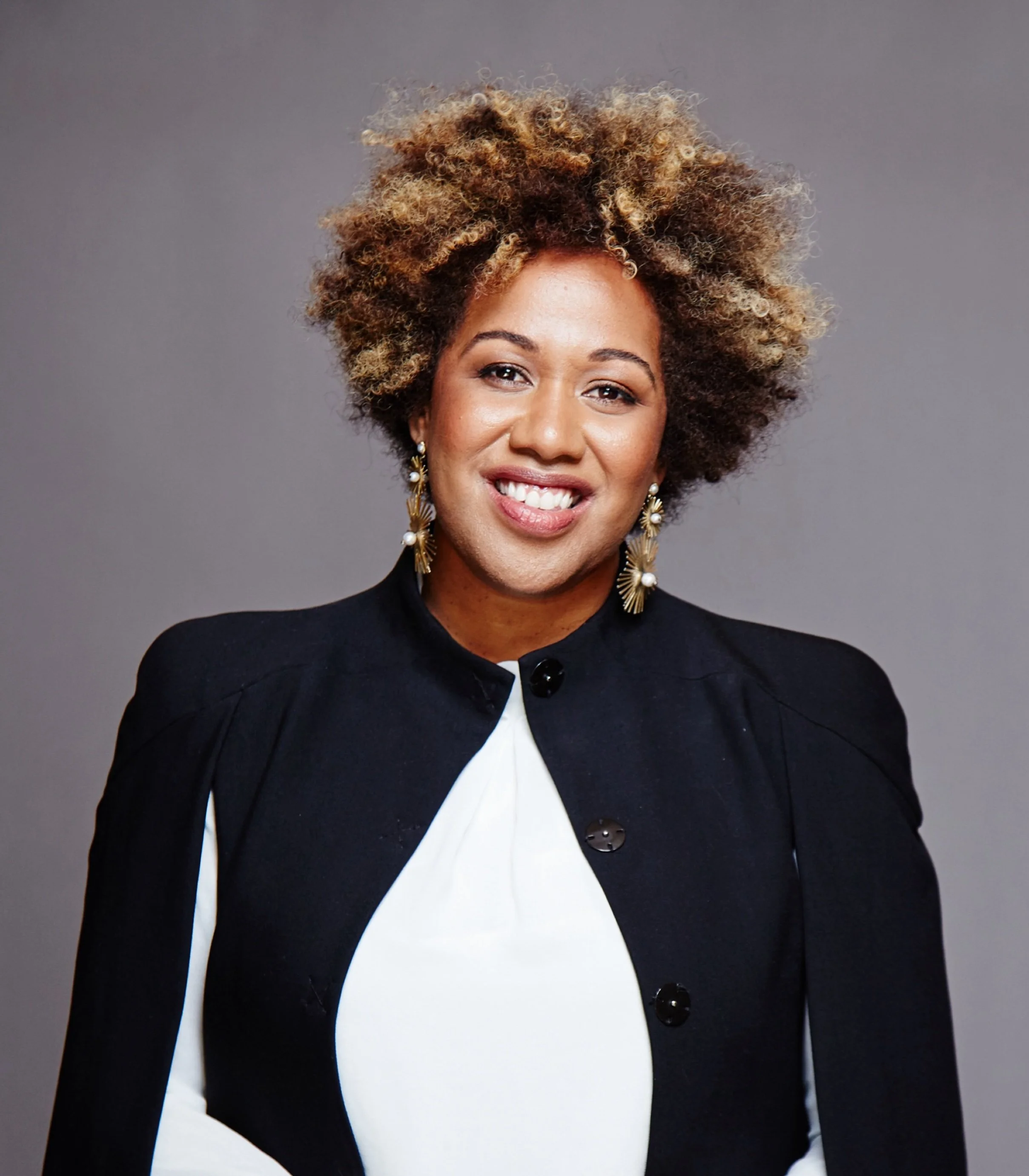 Portrait of a woman with curly blonde and brown hair, wearing a black blazer with large black buttons, a white blouse, and gold earrings with pearls, standing against a grey background. Elle McCarthy, Kalokalo Travel