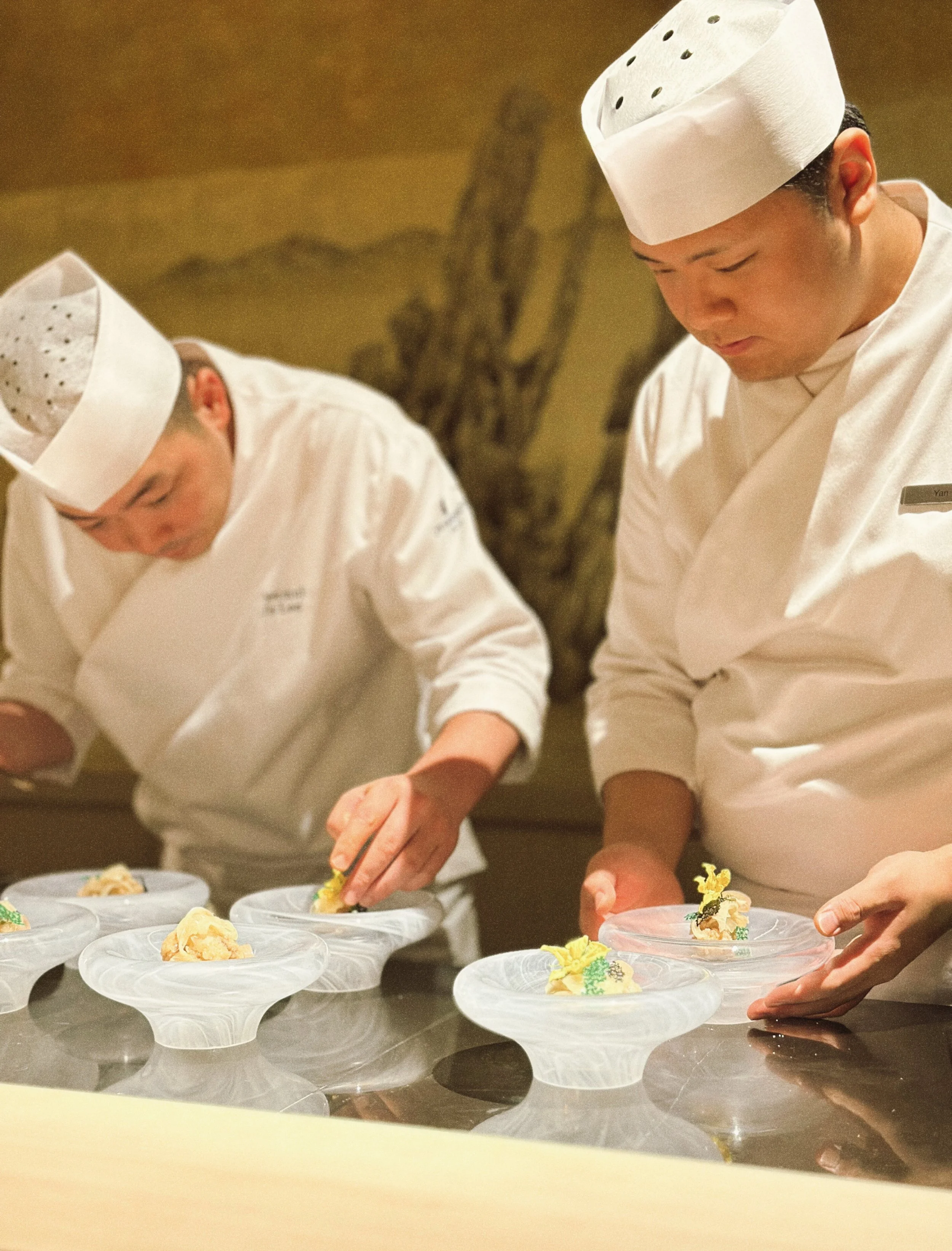 Two chefs in white uniforms and hats working together in a professional kitchen, preparing decorated dishes on plates.