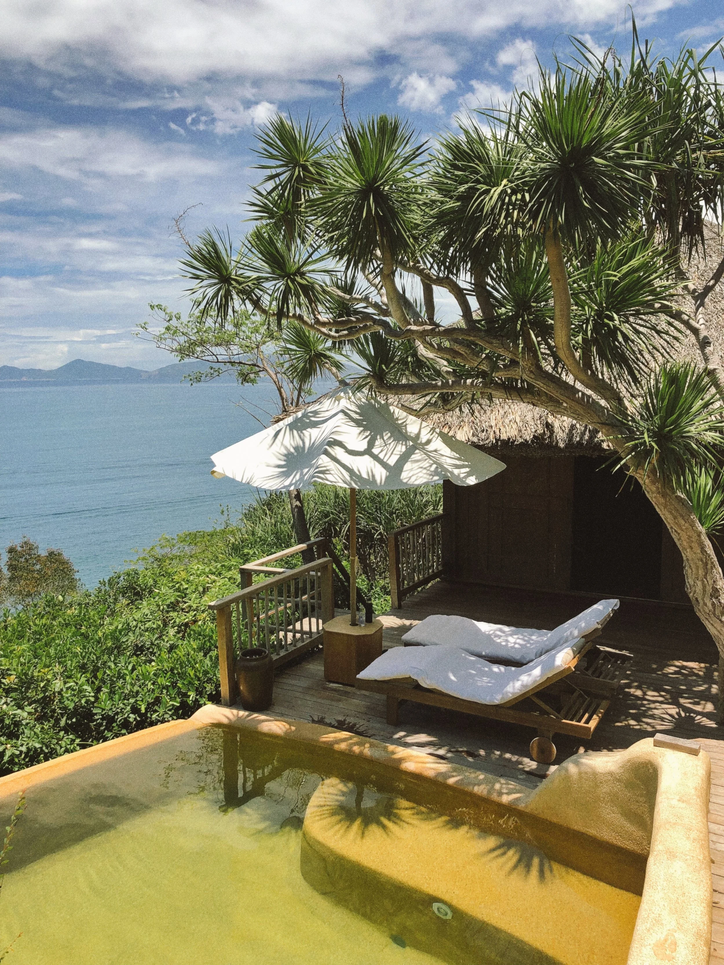 A patio area overlooking a body of water with mountains in the background. There is a large tropical tree with long, pointed leaves, a white umbrella, a wooden lounge chair with white cushions, and some greenery including bushes and grass. The sky is partly cloudy.