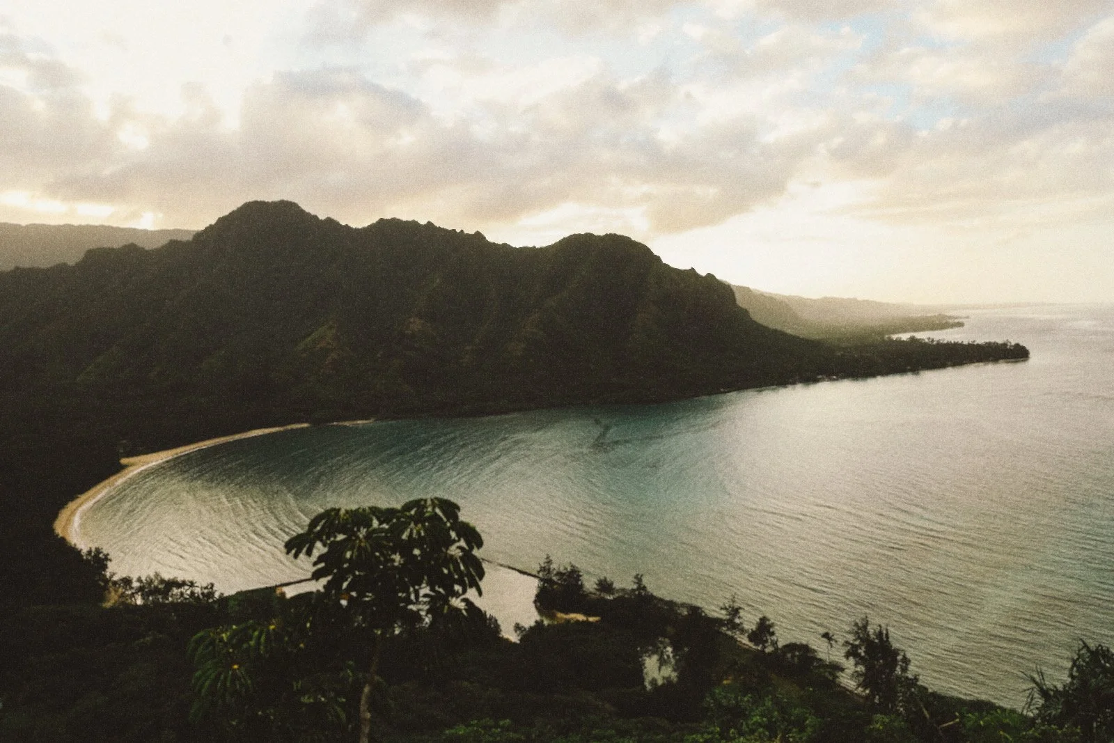 A coastal landscape with mountains in the background, a beach area, and calm waters with gentle ripples, under a cloudy sky.