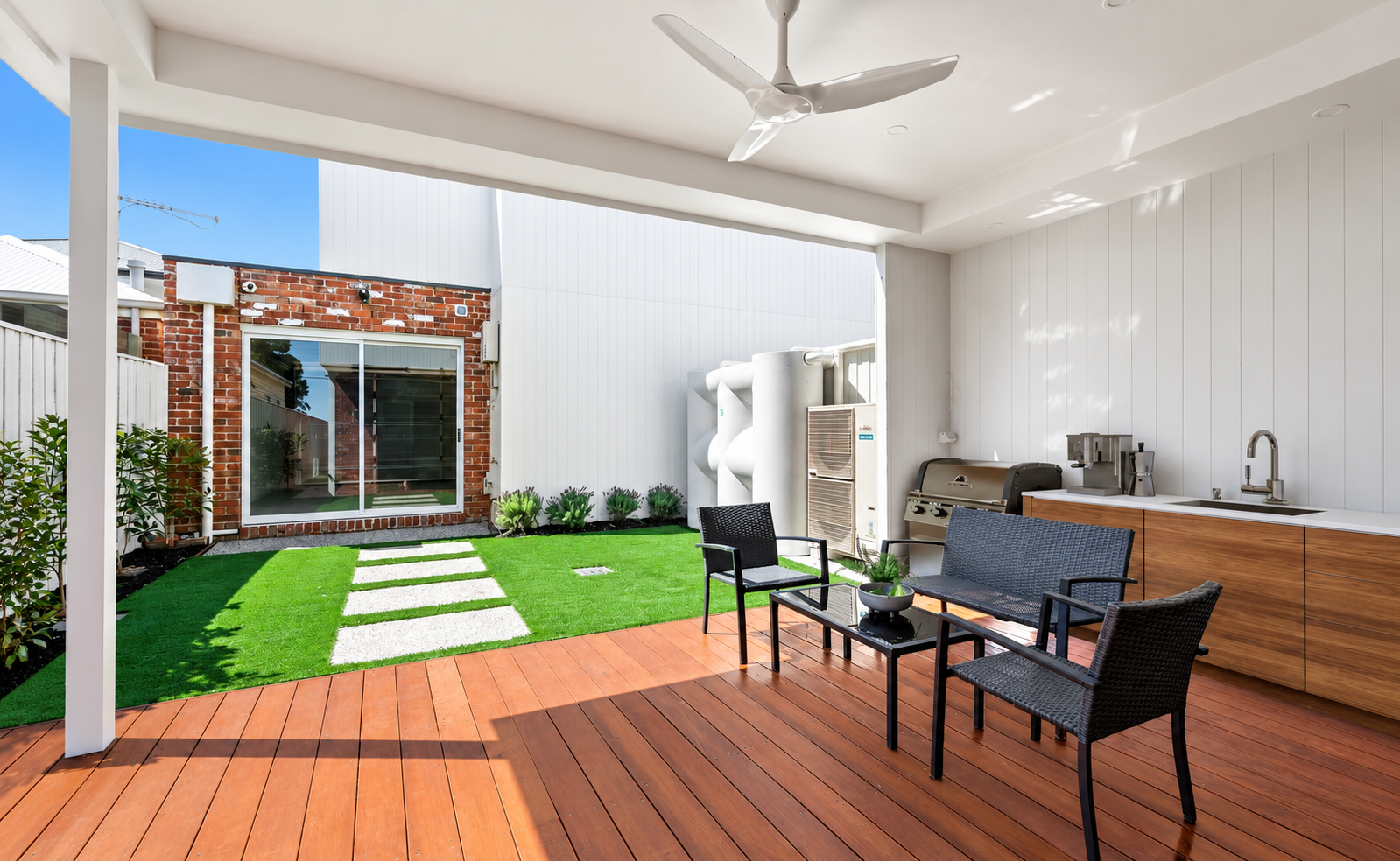 Covered outdoor patio with black patio furniture, a small gardening table, a stove, grill, sink, and storage cabinets, overlooking a small yard with green artificial turf, potted plants, and a brick building with glass door in the background.