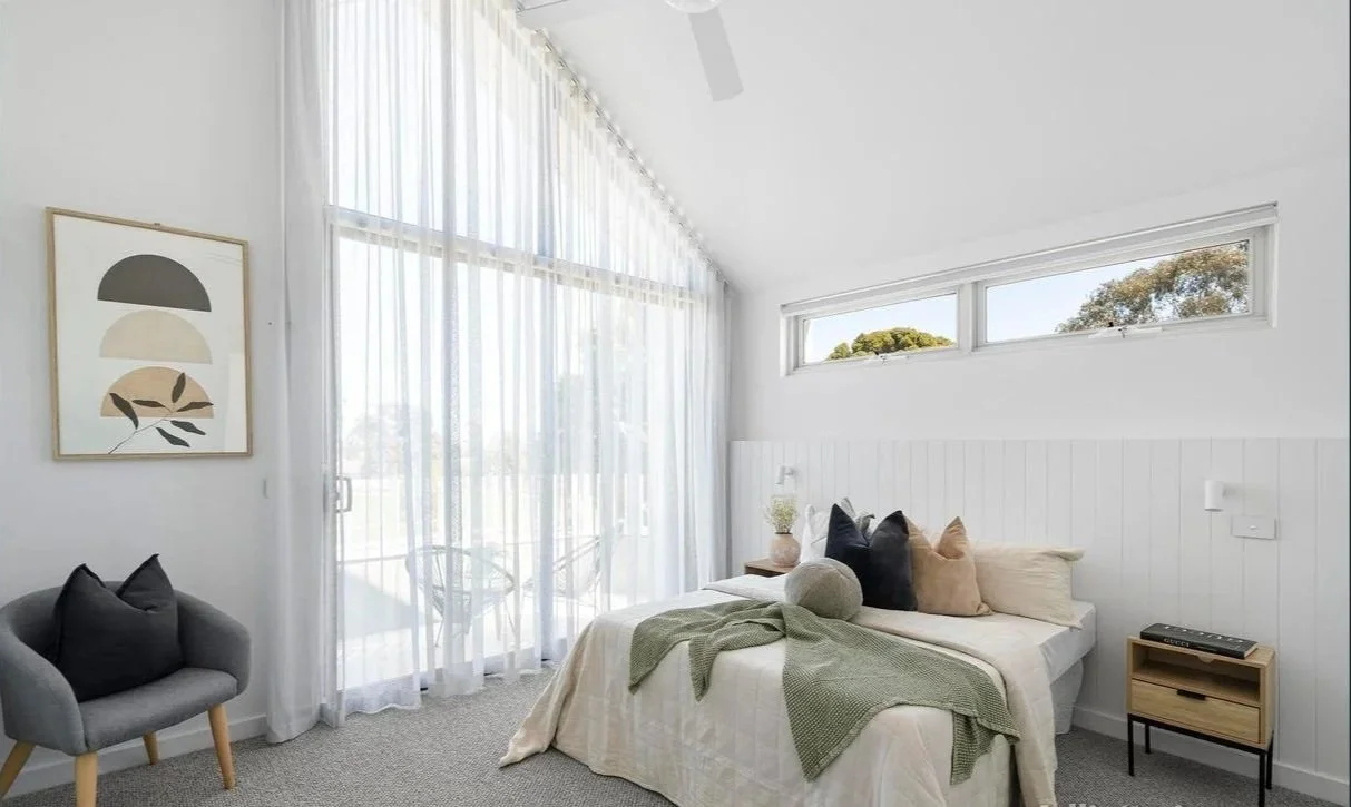 A bright bedroom with white walls and sheer curtains, a bed with neutral-colored pillows and a blanket, a gray armchair, a small side table with décor, and a large window with a view of trees.