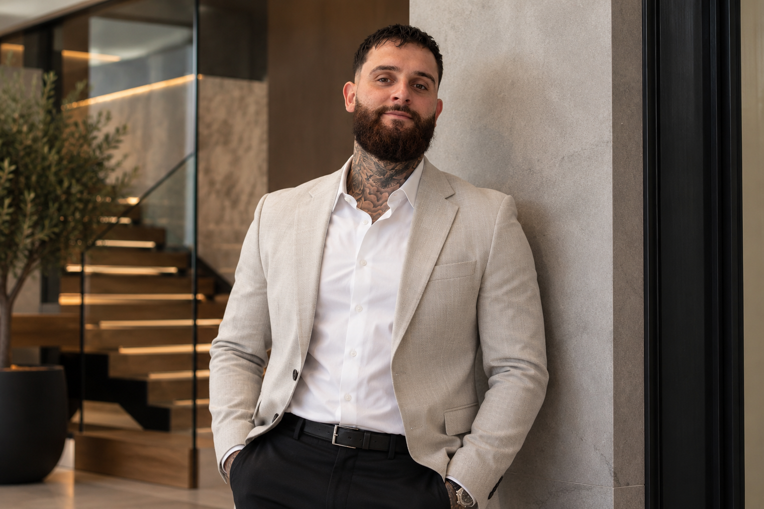 Man with beard and tattoos in a beige suit and white shirt, leaning against a wall in a modern building lobby.