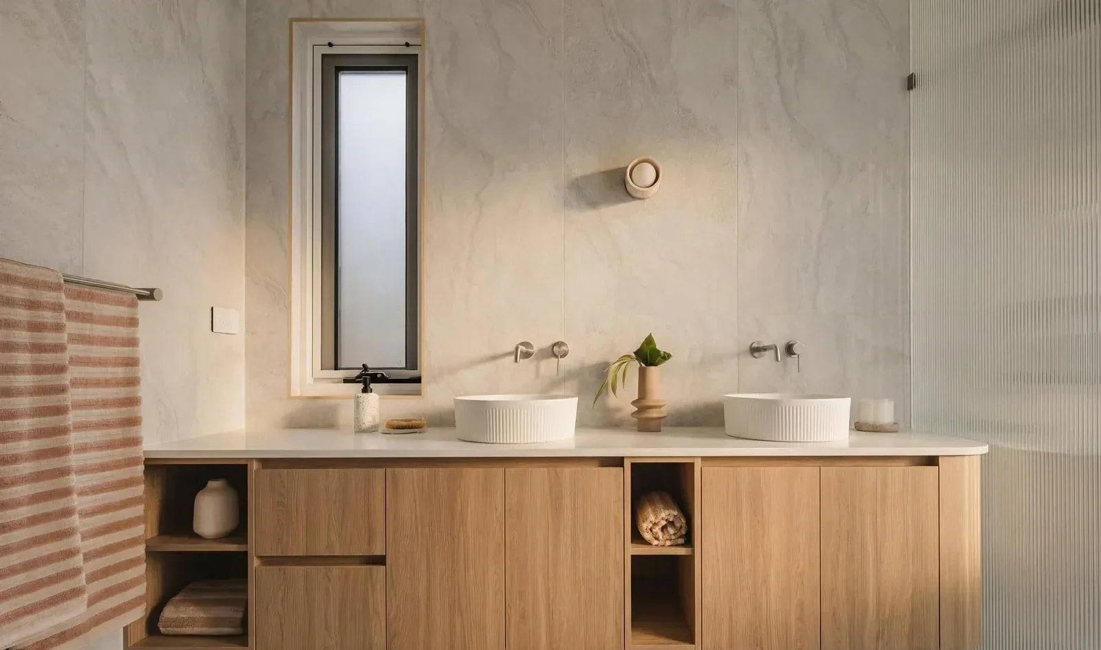 Bathroom vanity with two white vessel sinks, a wooden cabinet, a potted plant, soap dispenser, towels, and a window with frosted glass.