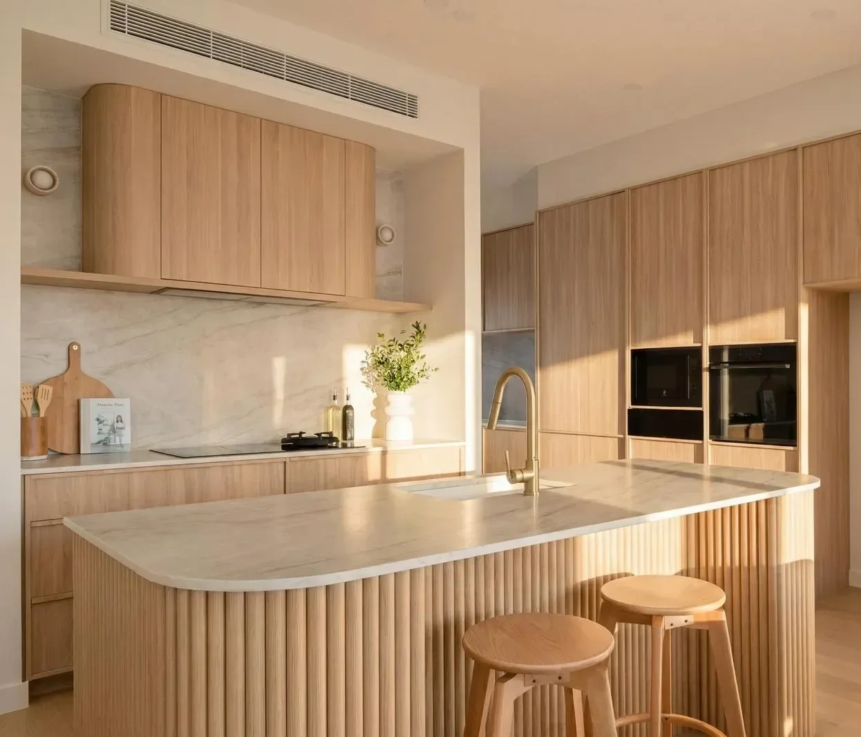 Modern kitchen with light wood cabinets, marble countertops, a central island with a rounded edge, two wooden stools, a potted plant, and built-in appliances including an oven and microwave.