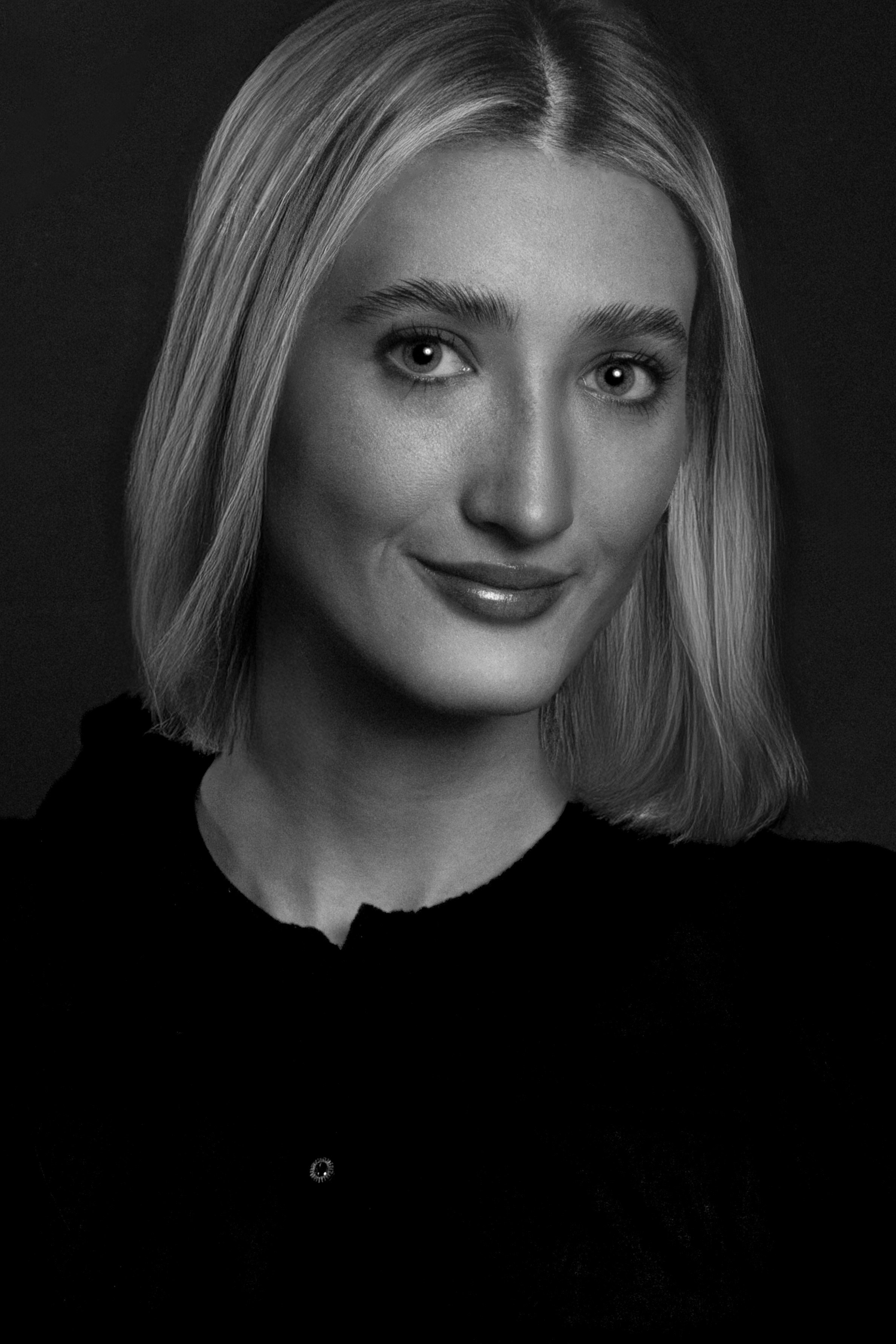 Black and white portrait of a young woman with shoulder-length hair, smiling slightly, wearing a dark top, against a dark background.