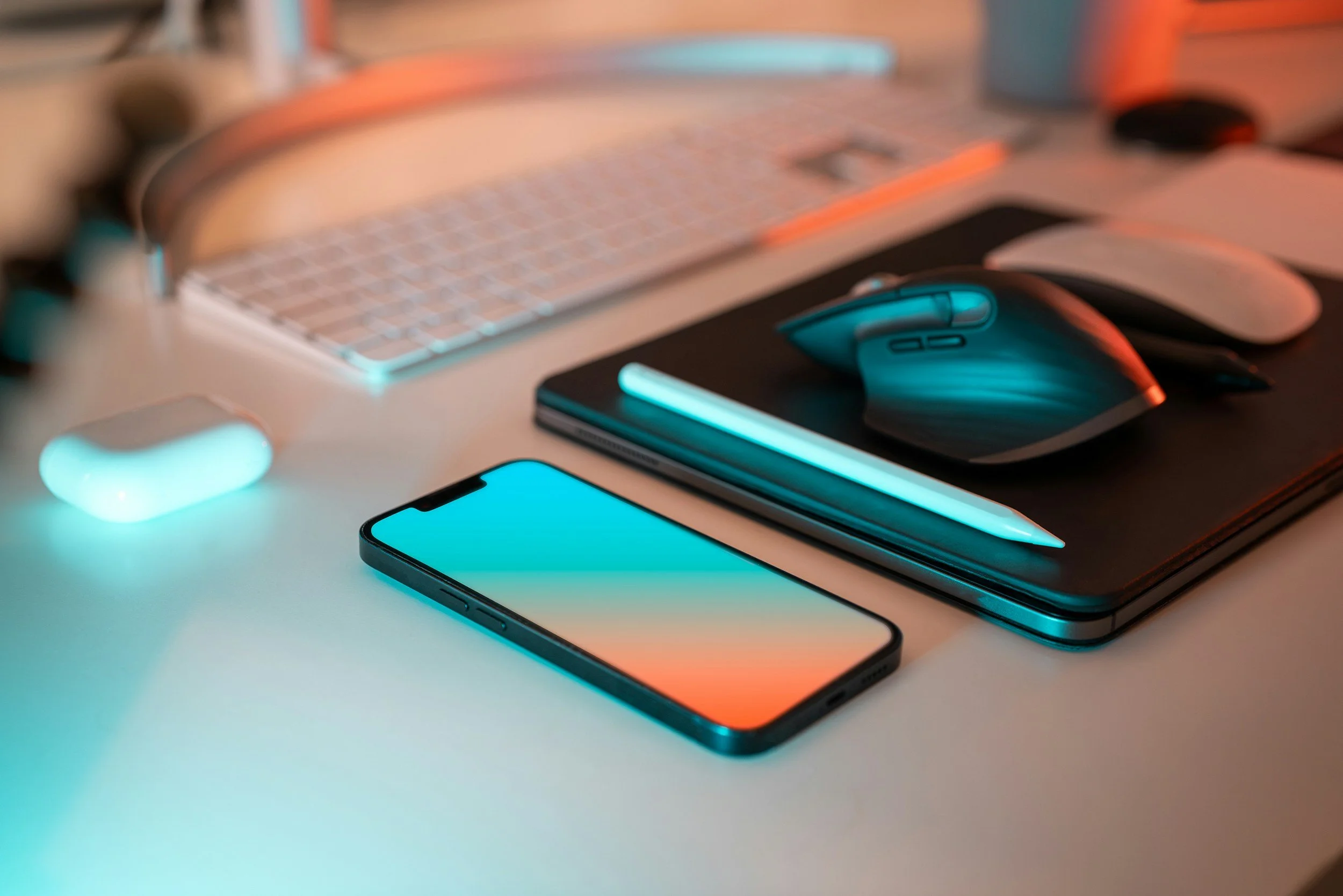 A workspace with a smartphone, wireless earbuds, a mouse, a keyboard, a tablet, and a laptop on a desk, illuminated by soft teal and orange lighting.