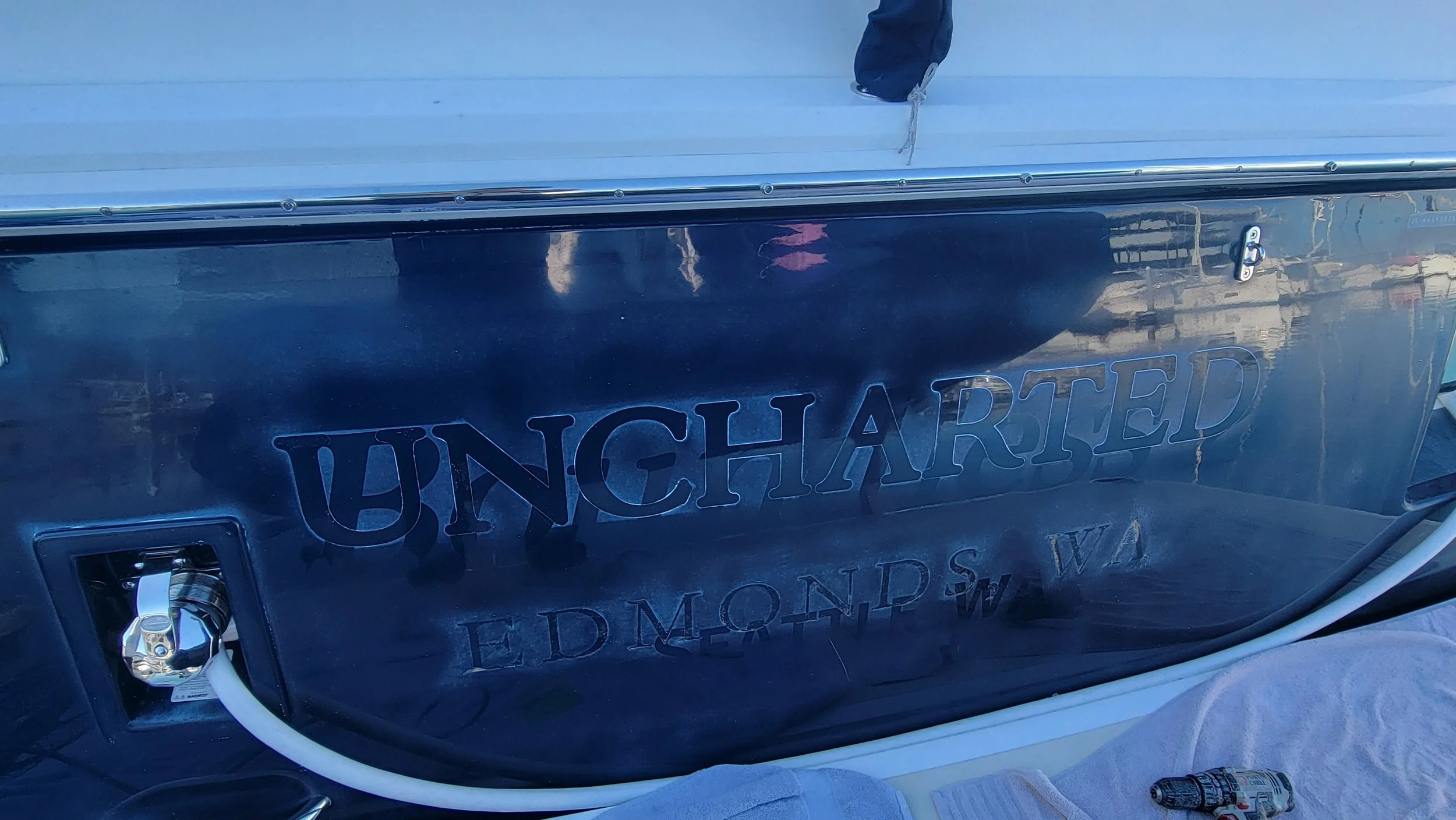 Close-up of a dark blue boat hull labeled “UNCHARTED” and “EDMONDS, WA,” with chrome hardware and a white hose.