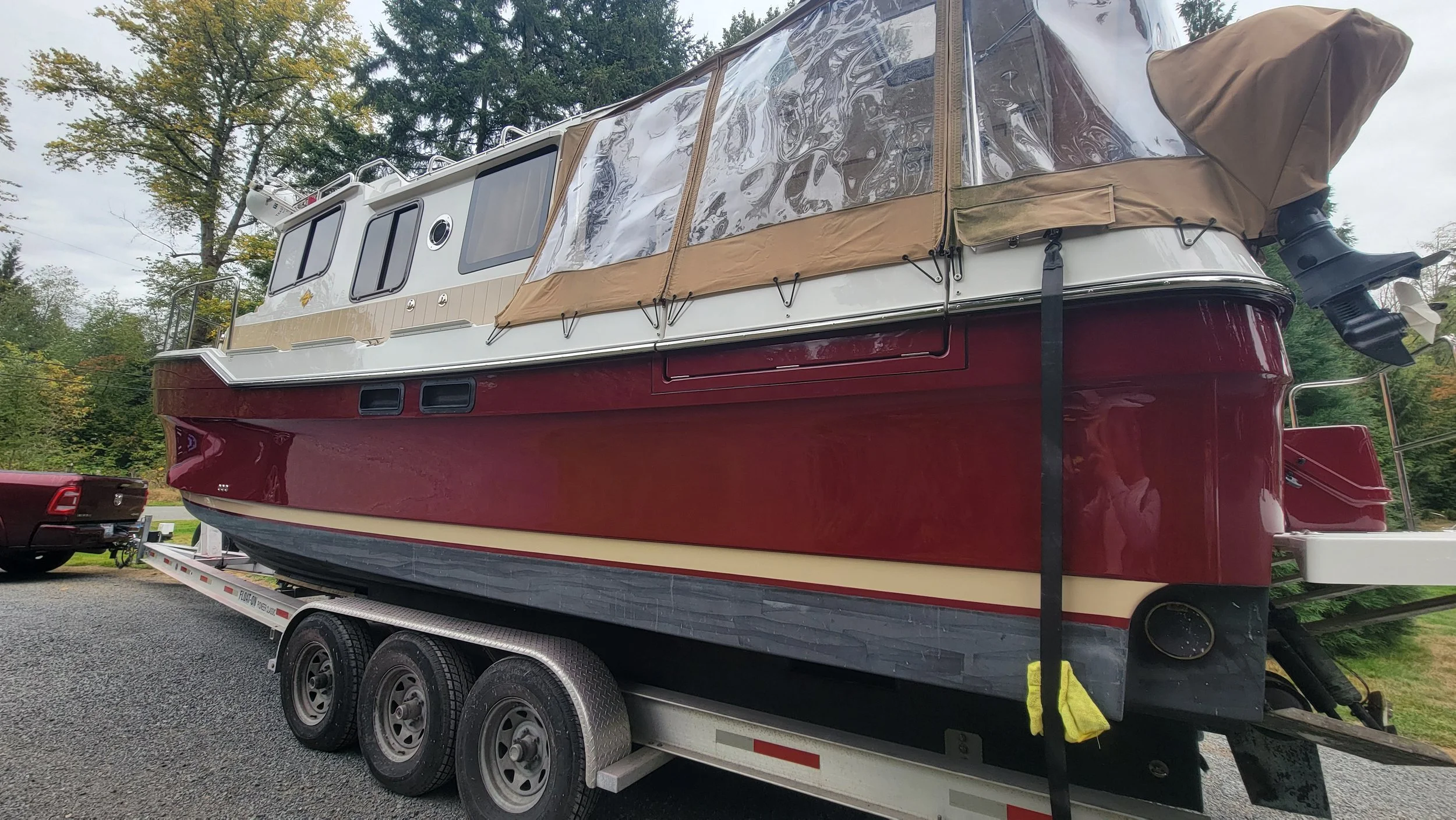 Large maroon-and-white cabin cruiser boat with tan canvas cover on a triple-axle trailer outdoors
