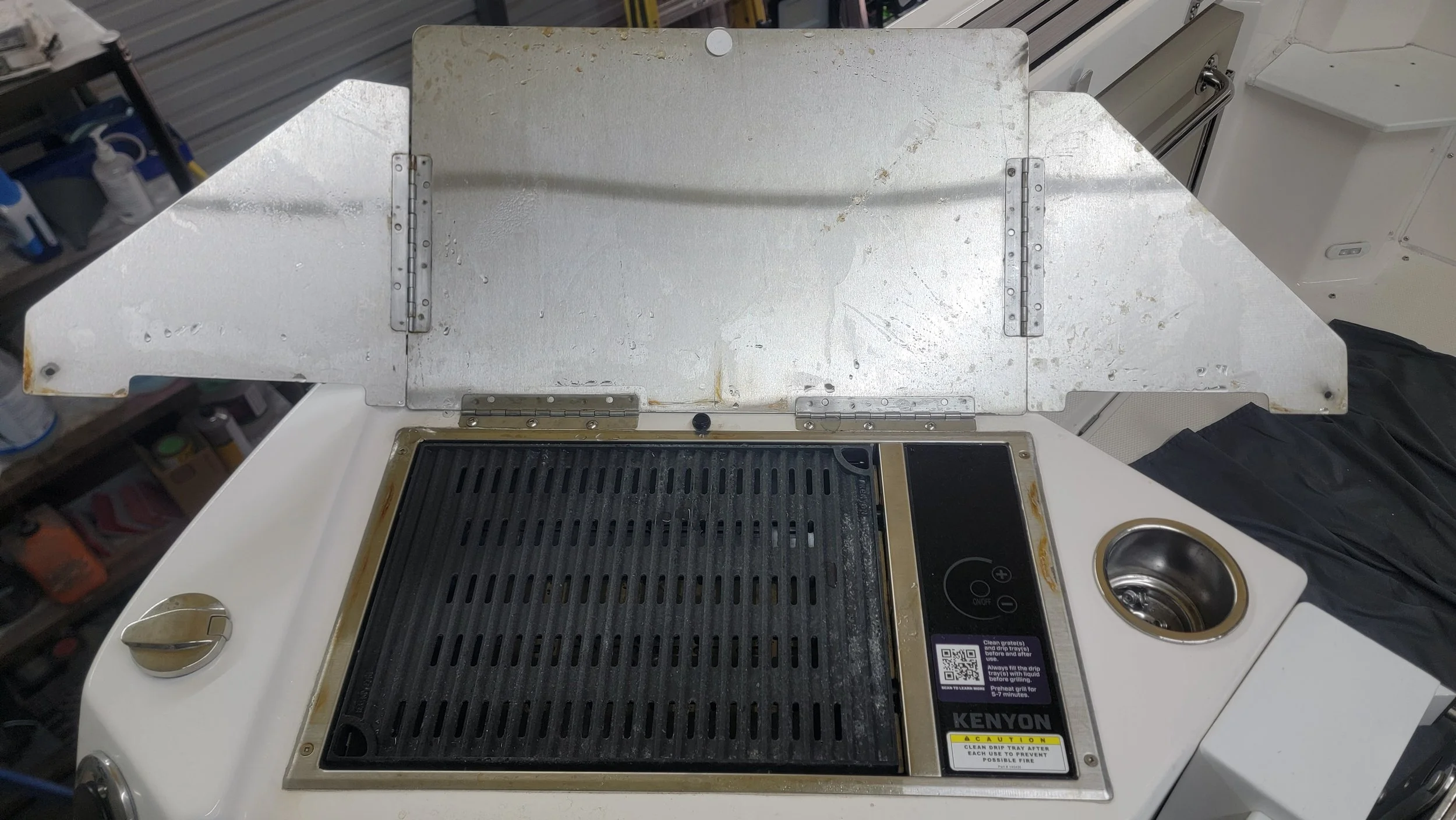 Kenyon electric marine grill with open metal lid, black grate, control panel, and adjacent stainless-steel sink on a boat
