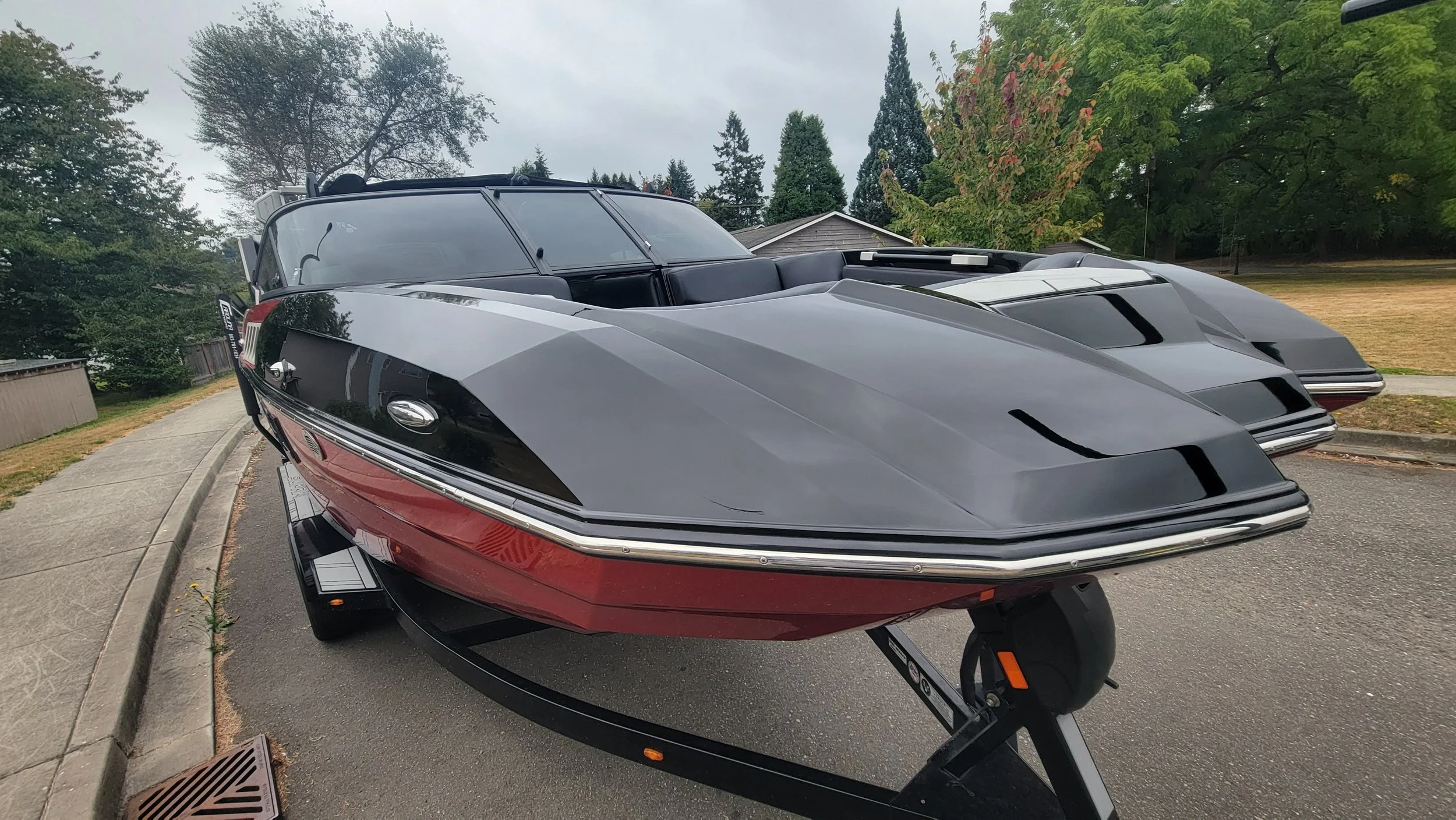 Black, gray, and red wakeboard boat on a trailer parked beside a residential sidewalk.