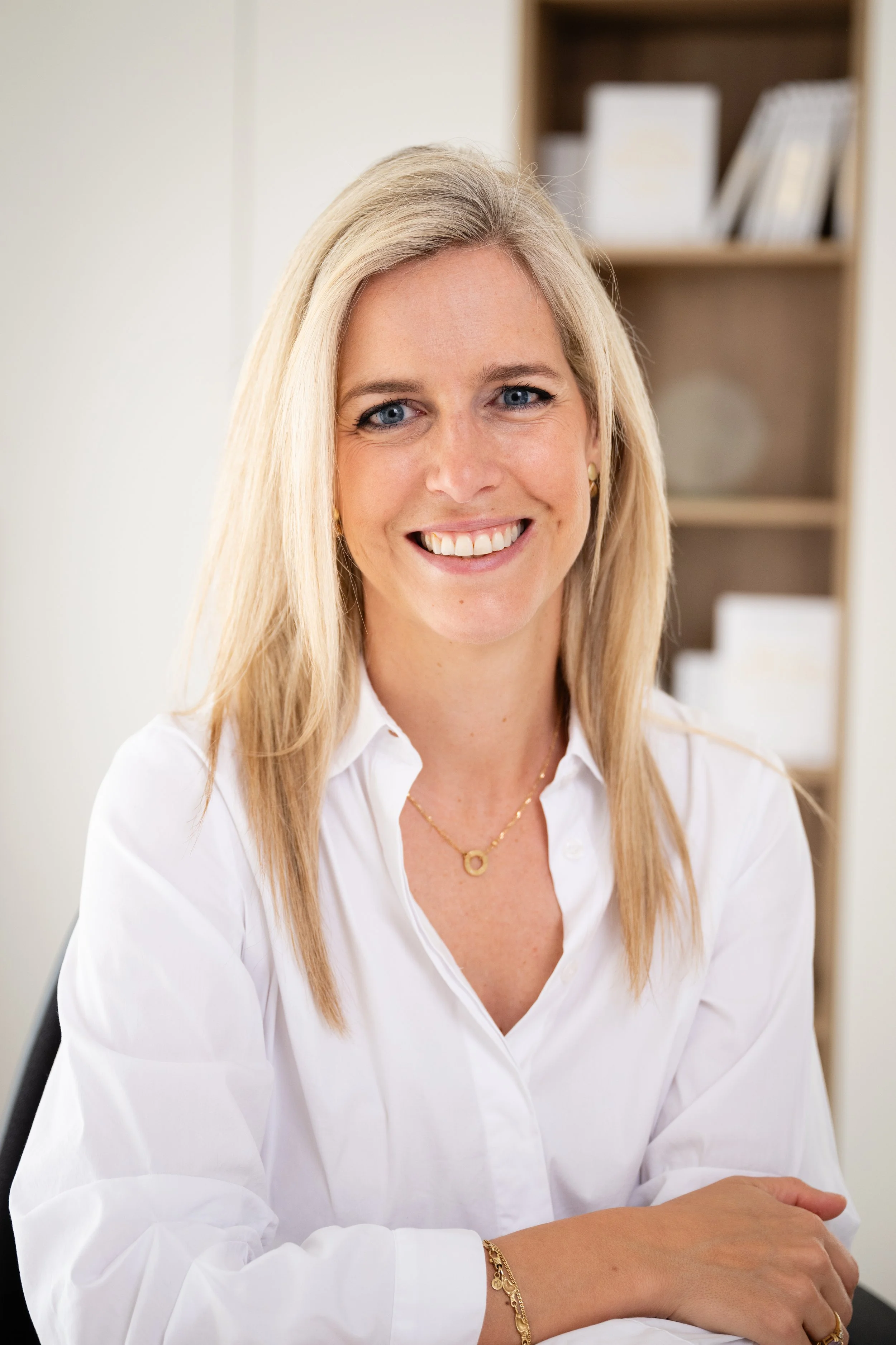 Close-up of a smiling blonde woman wearing a white blouse, gold jewelry, and sitting at a desk in an office.