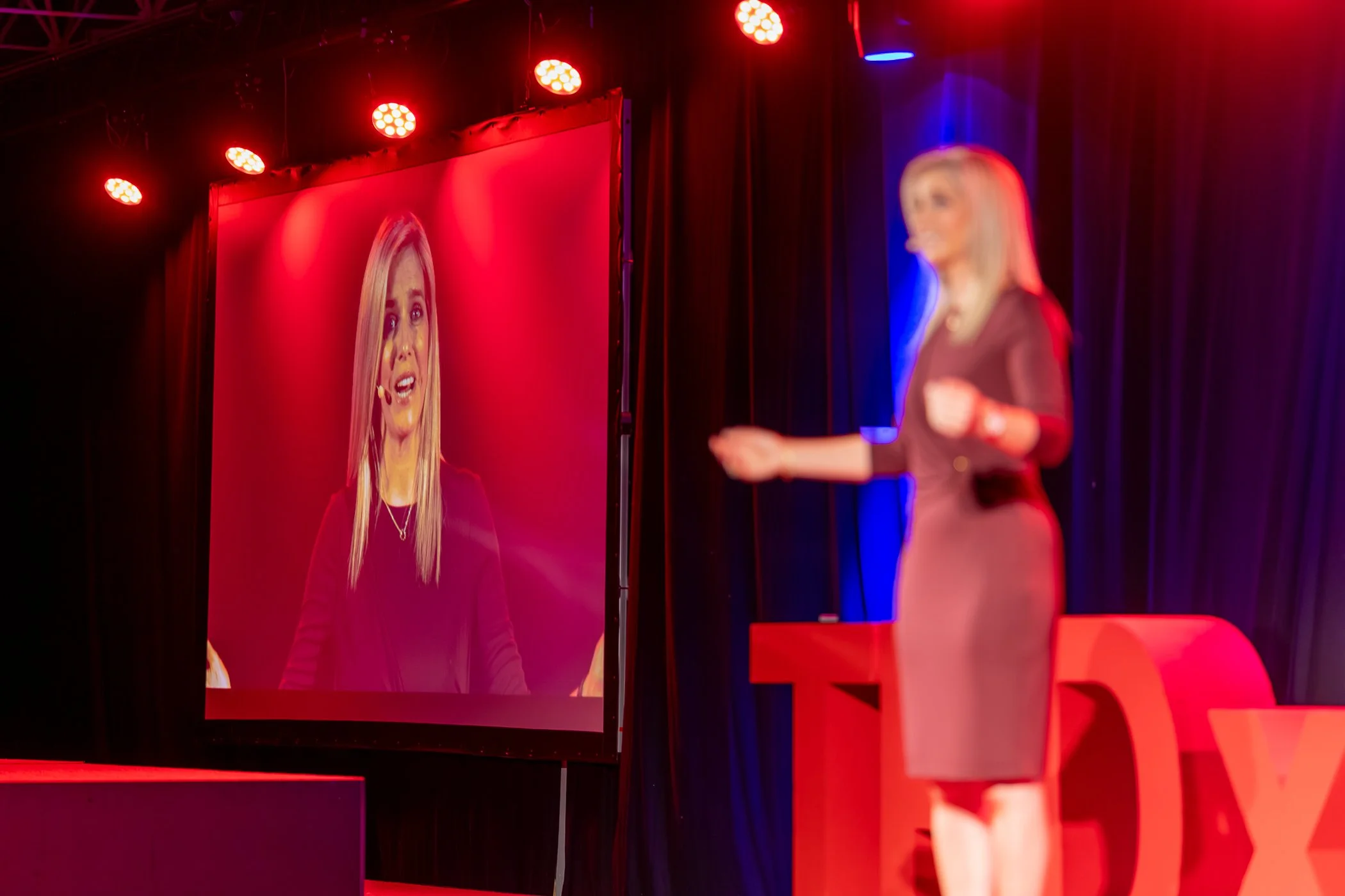 A woman on stage speaking at a conference or presentation, with a large screen behind her displaying her image.