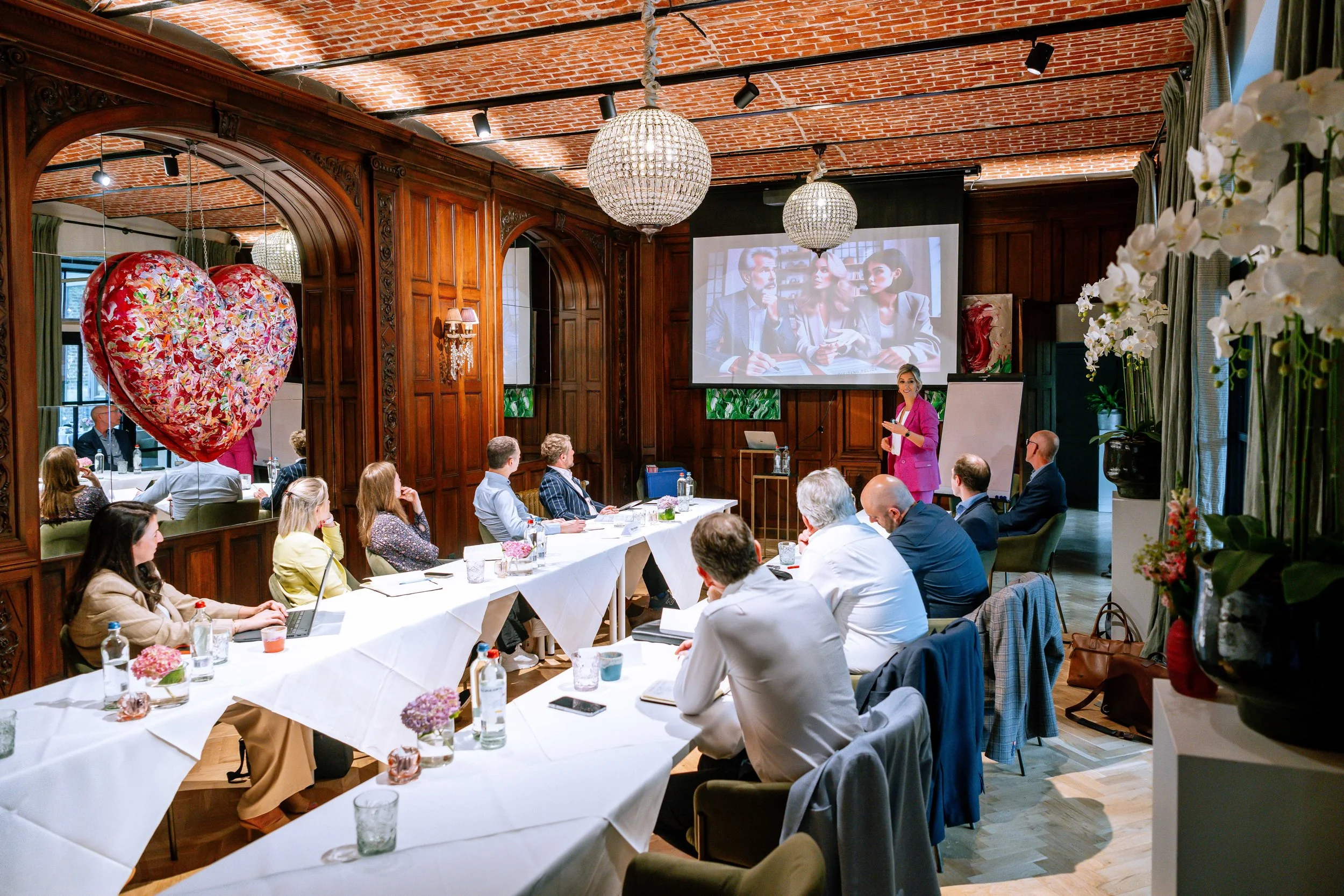 A conference room filled with people seated at long tables, listening to a woman in a pink blazer giving a presentation. There are large chandeliers hanging from a brick ceiling, and a big screen displays a slide with three people in conversation. The room has wooden paneling and is decorated with flowers and large potted plants.
