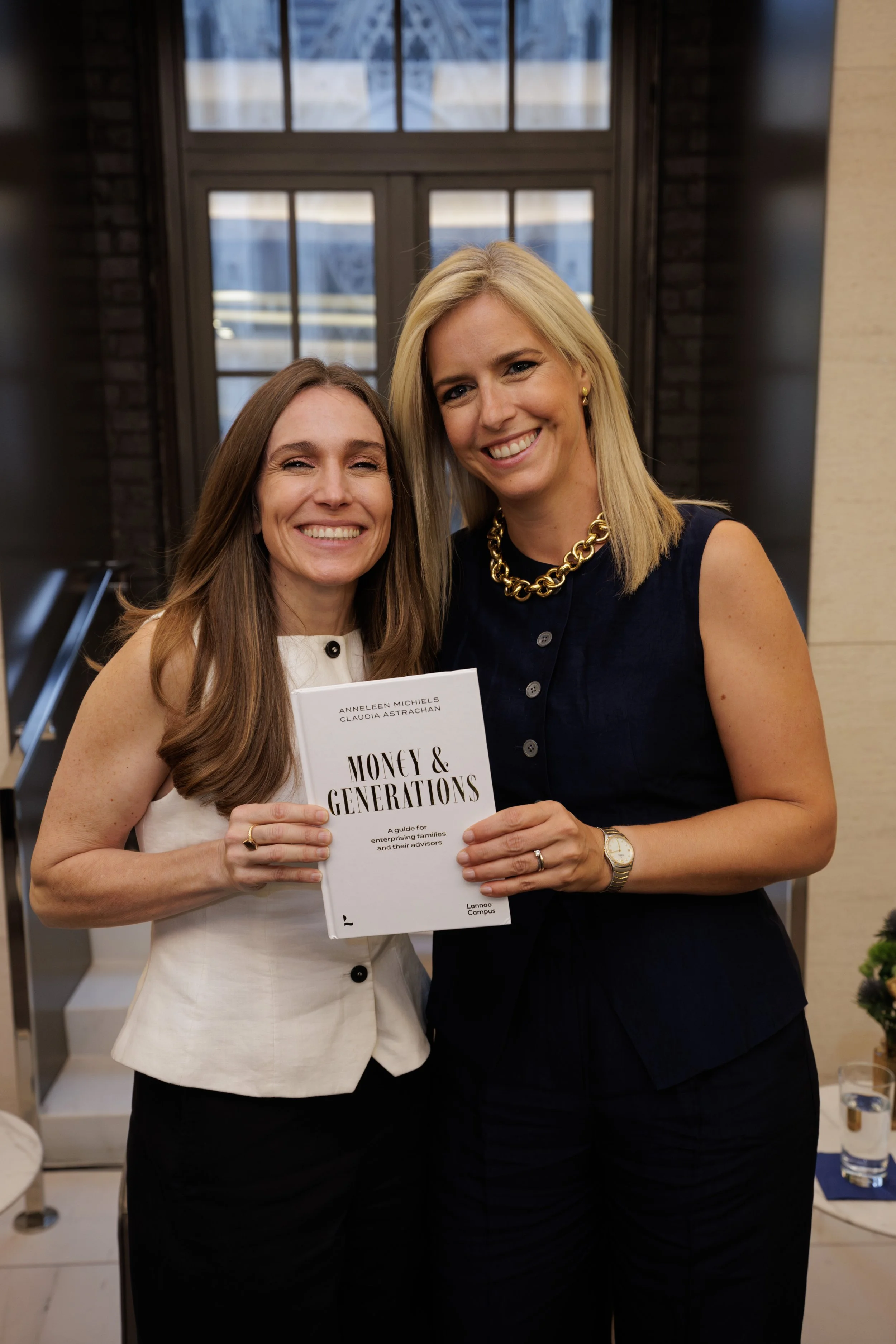 Two women smiling and standing close together indoors, holding a book titled 'Money & Generations.' One woman has long brown hair and is wearing a sleeveless white top, while the other woman has blonde hair and is wearing a sleeveless dark blue top with a gold chain necklace. The background features large windows and an indoor setting.