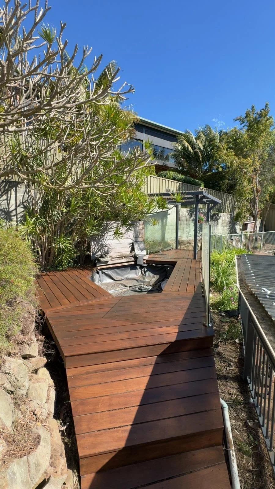 Wooden backyard deck with a covered seating area, tropical plants, and a modern house under a clear blue sky