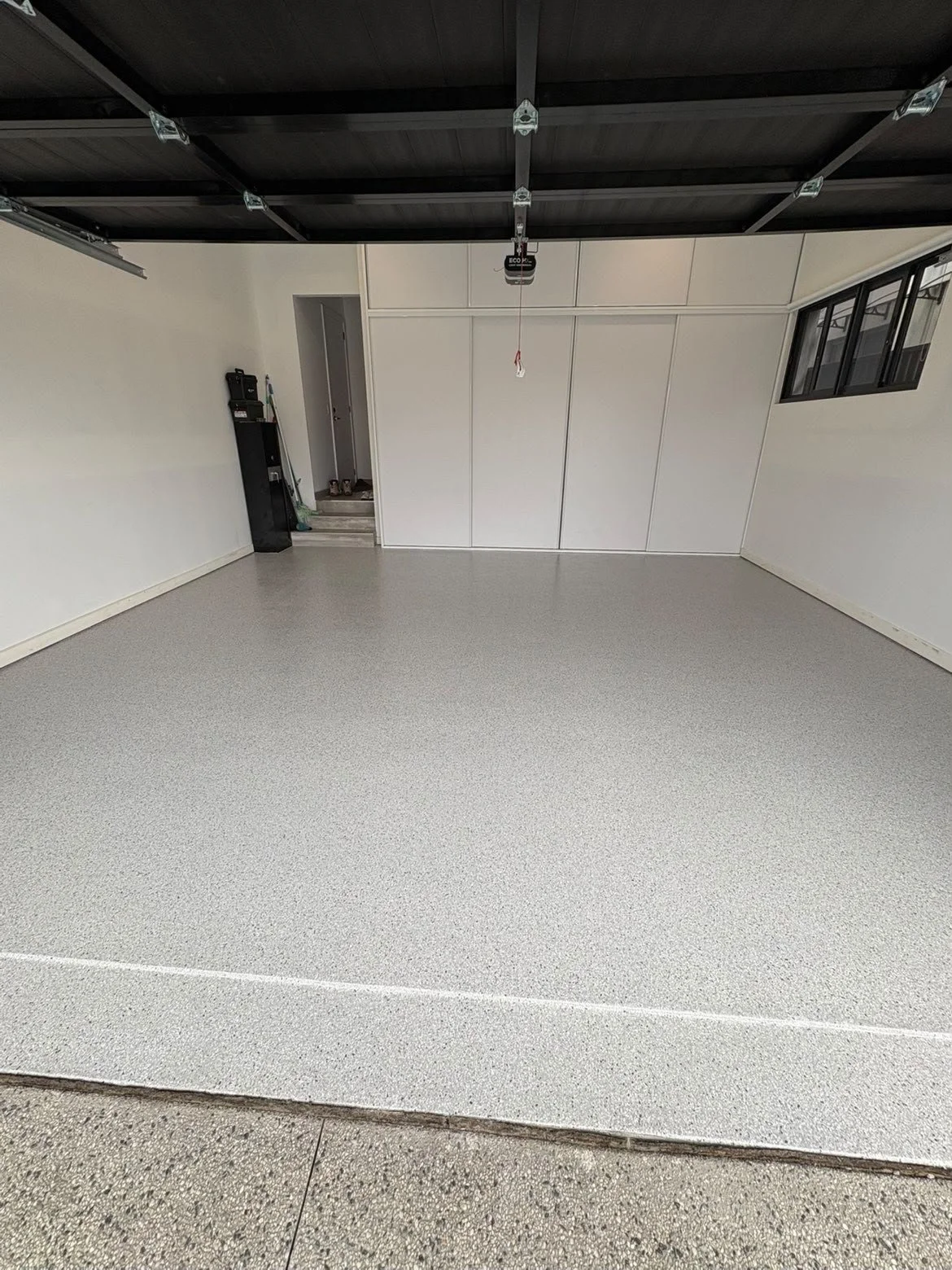 Clean, empty residential garage with a speckled epoxy floor, open garage door, white walls, storage cabinets, and windows.